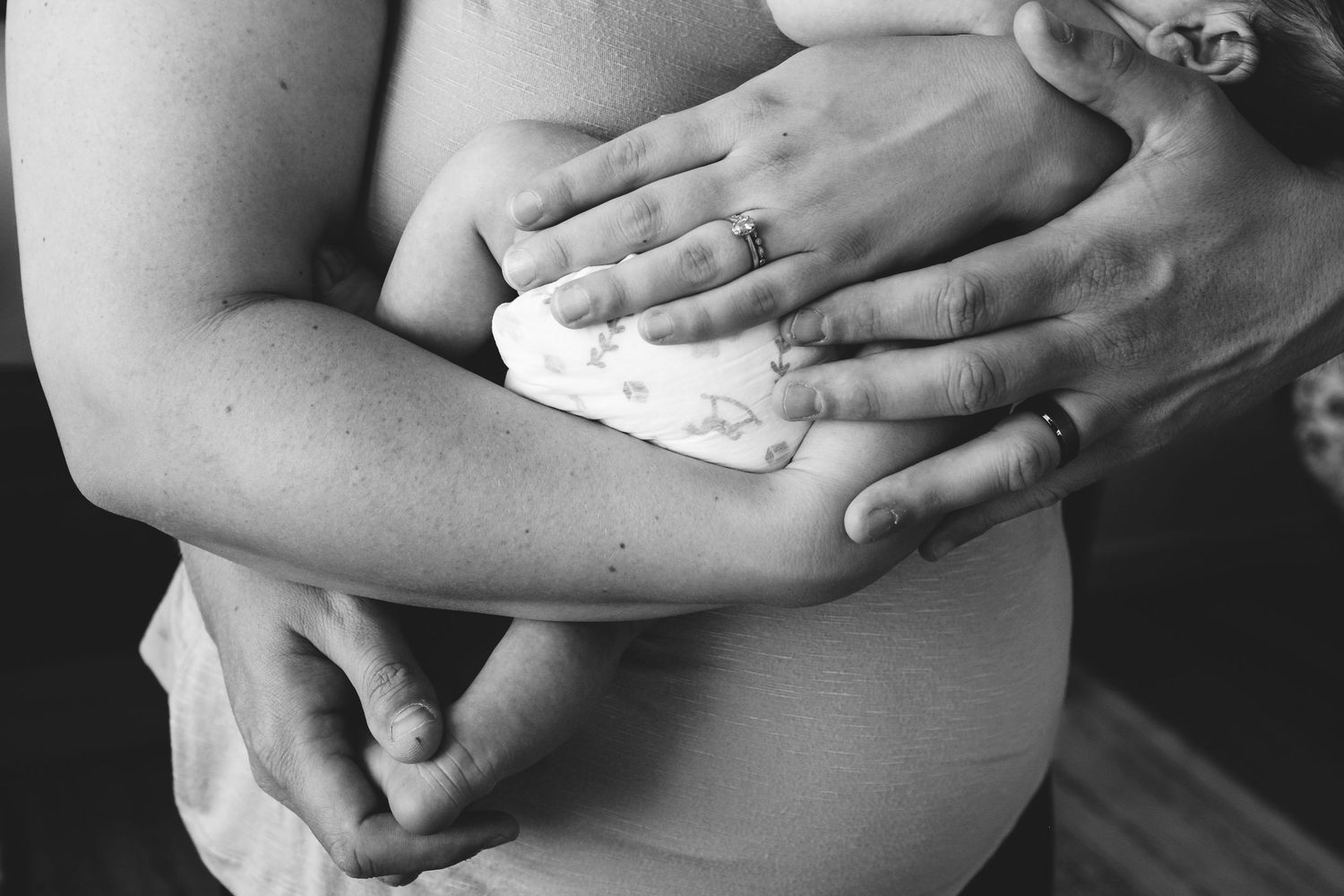 Black and white close up of mother and father's hands with wedding rings as they snuggle newborn during Denver in-home photo session.