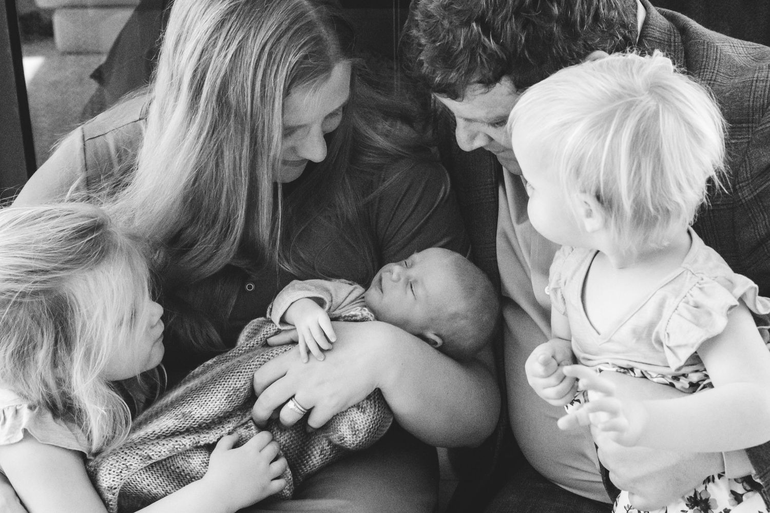 Mother holding newborn son, surrounded by father and two toddler daughters, all gazing lovingly at the baby.