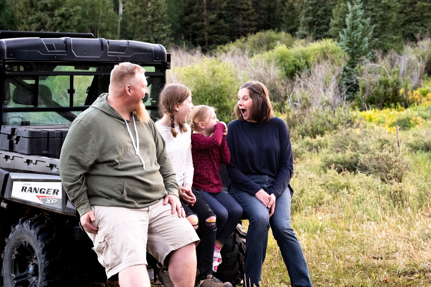Father, two daughters and mother sitting on the back of a UTV in a green mountain meadow, all laughing.