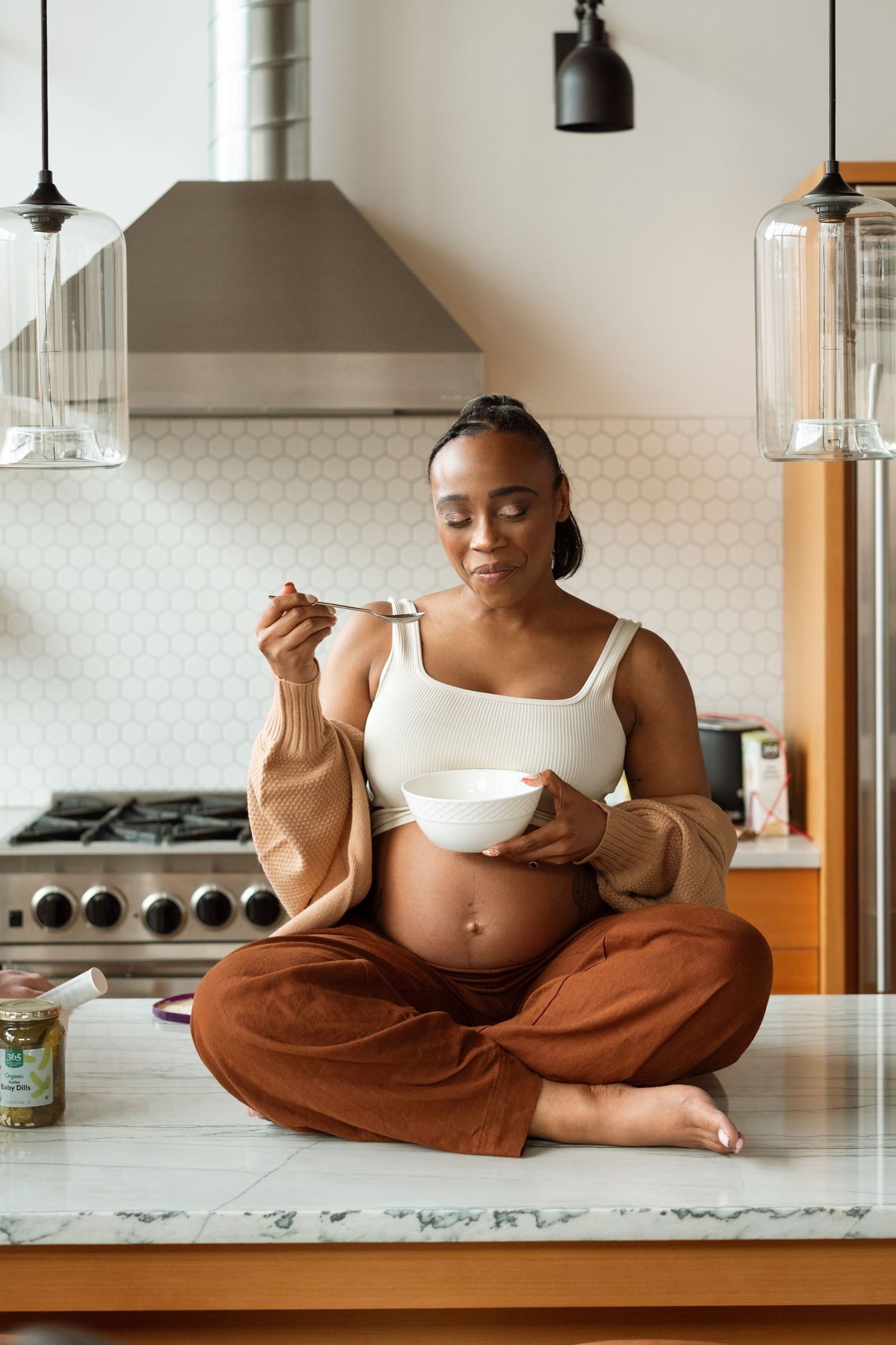 Pregnant mom sitting on counter in modern kitchen, eating ice cream.