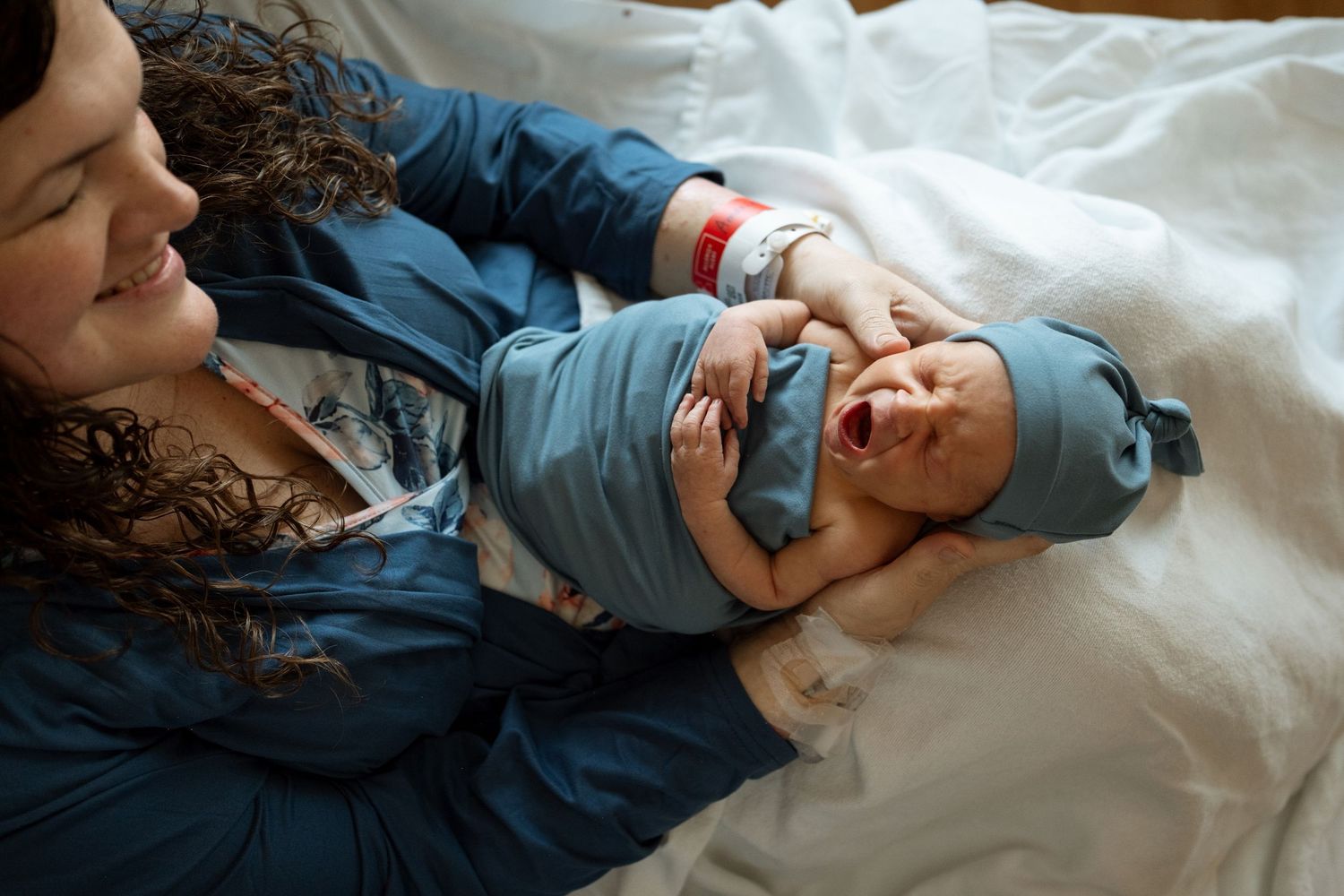 Mother sitting in hospital bed, holding newborn boy, swaddled in blue blanket, baby yawning.