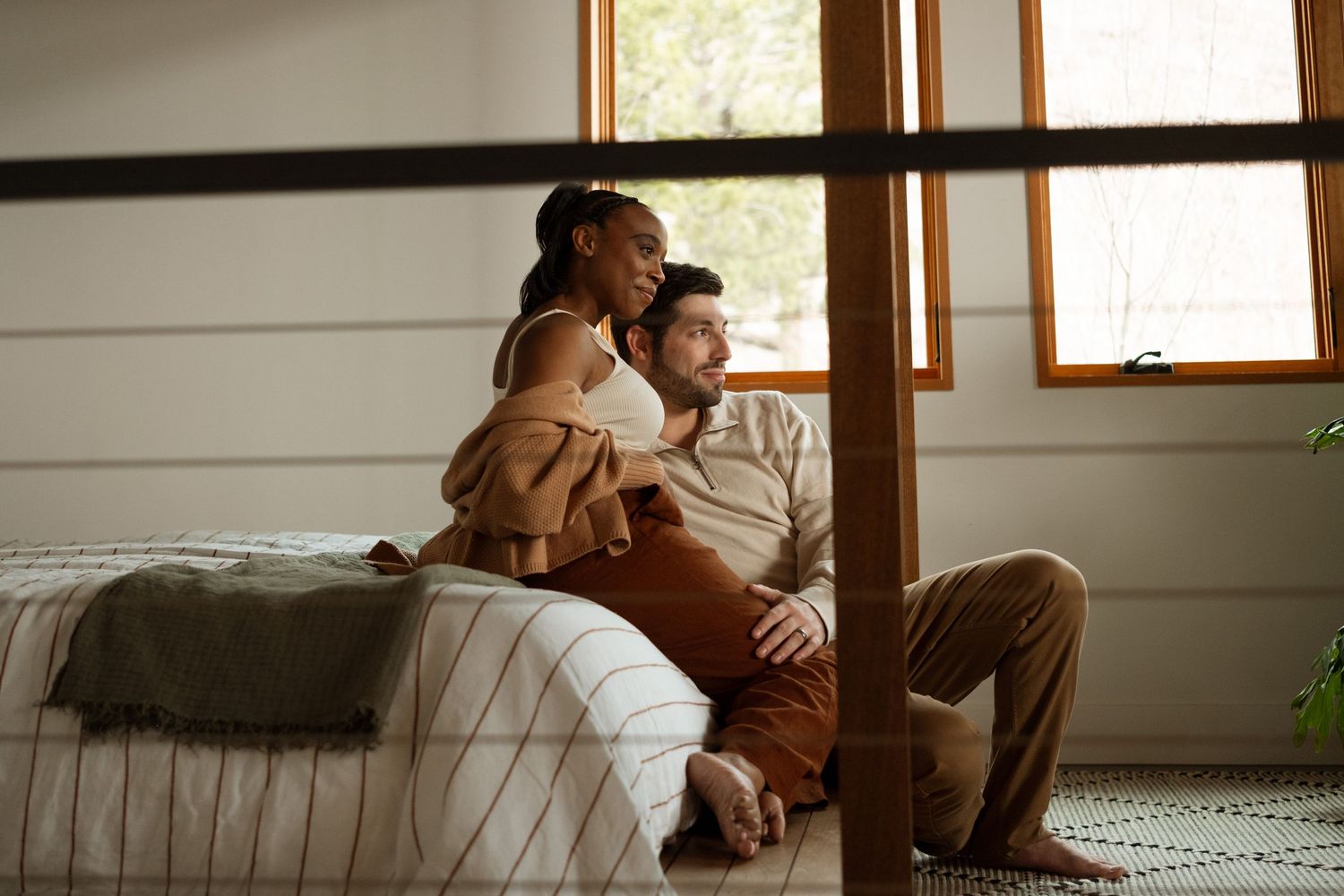 Pregnant couple sitting on bed behind modern staircase during maternity photography session near Denver, Colorado.