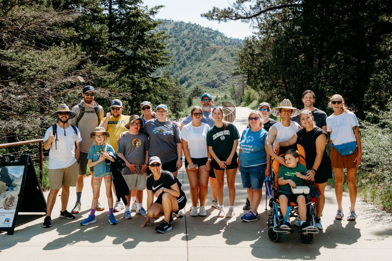 Posed group photo of WILD's hiking participants at Clear Creek Trail, standing on paved path, large evergreens in background.