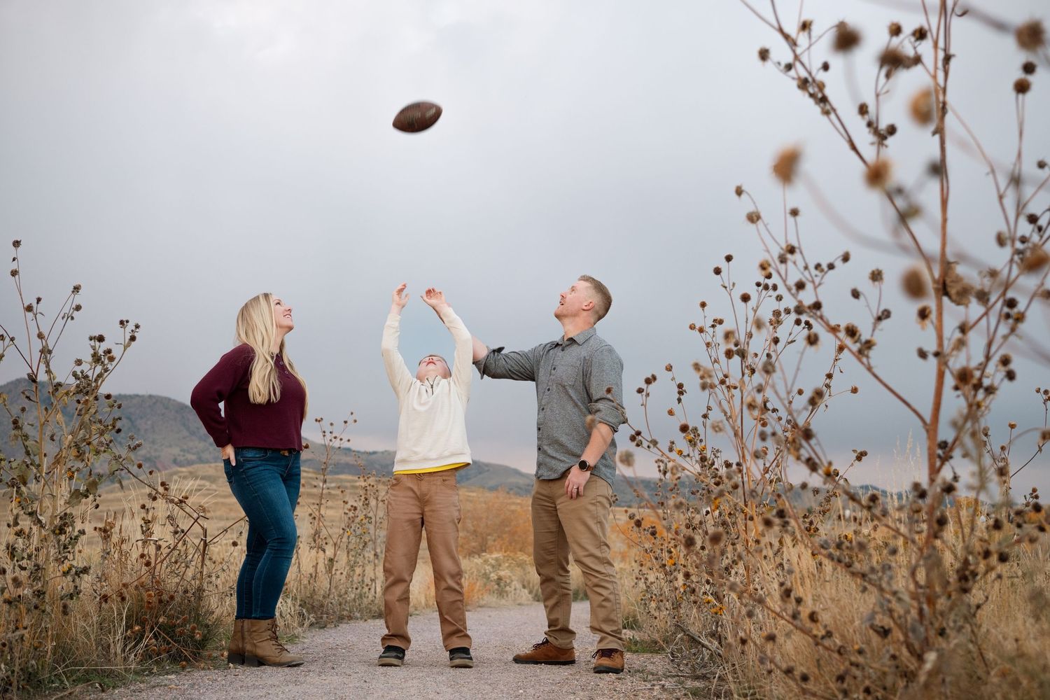 Family of three playfully throwing football in the air, surrounded by dried sunflowers.