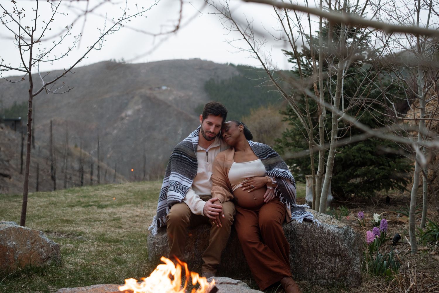 Pregnant couple snuggling outside, wrapped in blanket, campfire in the background.