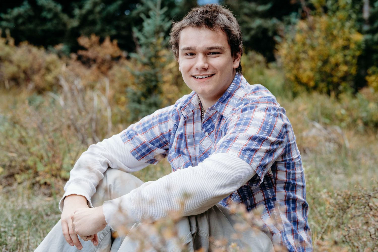 Senior portrait of teen boy sitting in the mountains, smiling, green evergreens in the background.