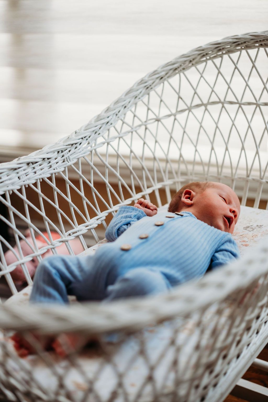 Newborn boy dressed in blue onesie, lay sleeping in white bassinet.
