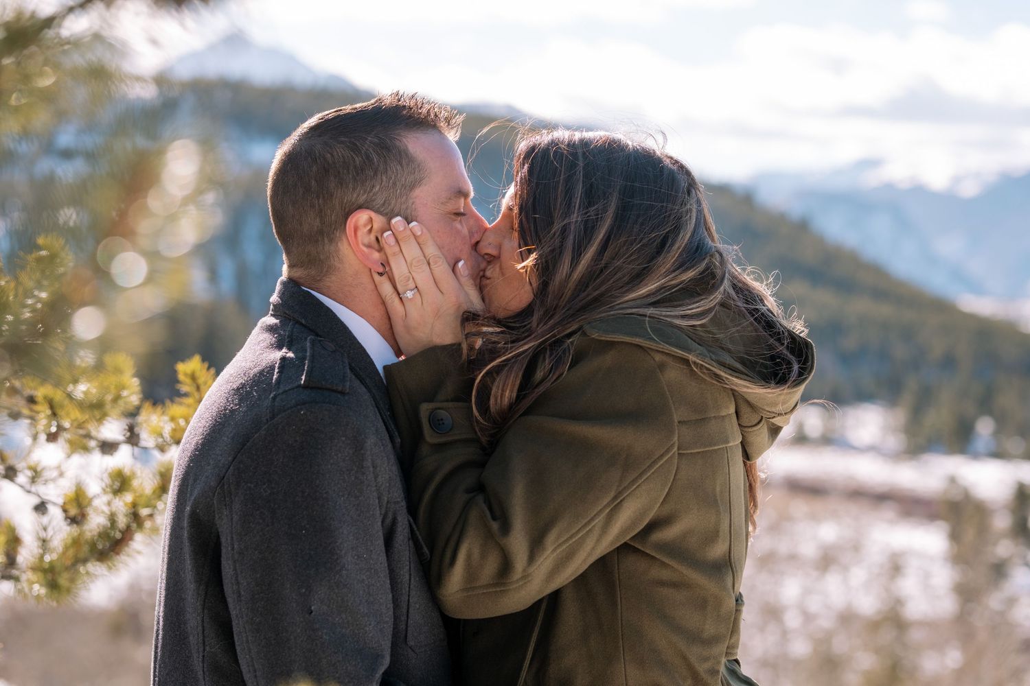Couple kissing during engagement photo session near Dillon, Colorado, mountains in the background, evergreen trees in foreground.