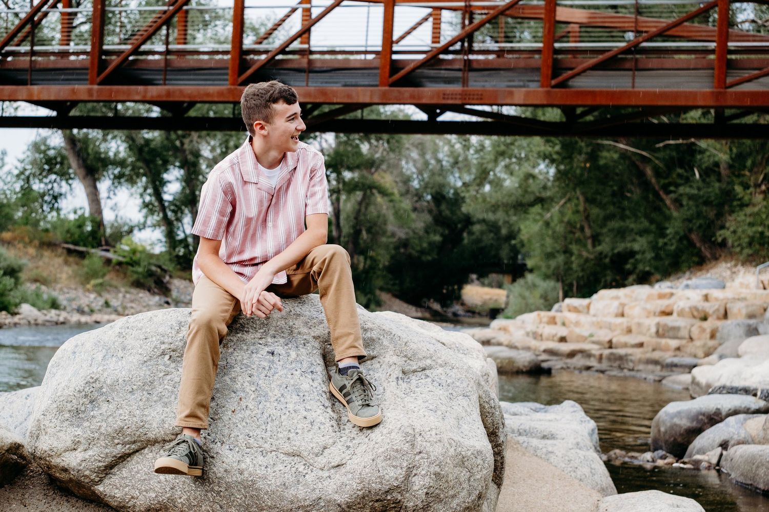 Senior portrait of teen boy sitting on a large boulder on the Cache la Poudre River with bridge in the background.