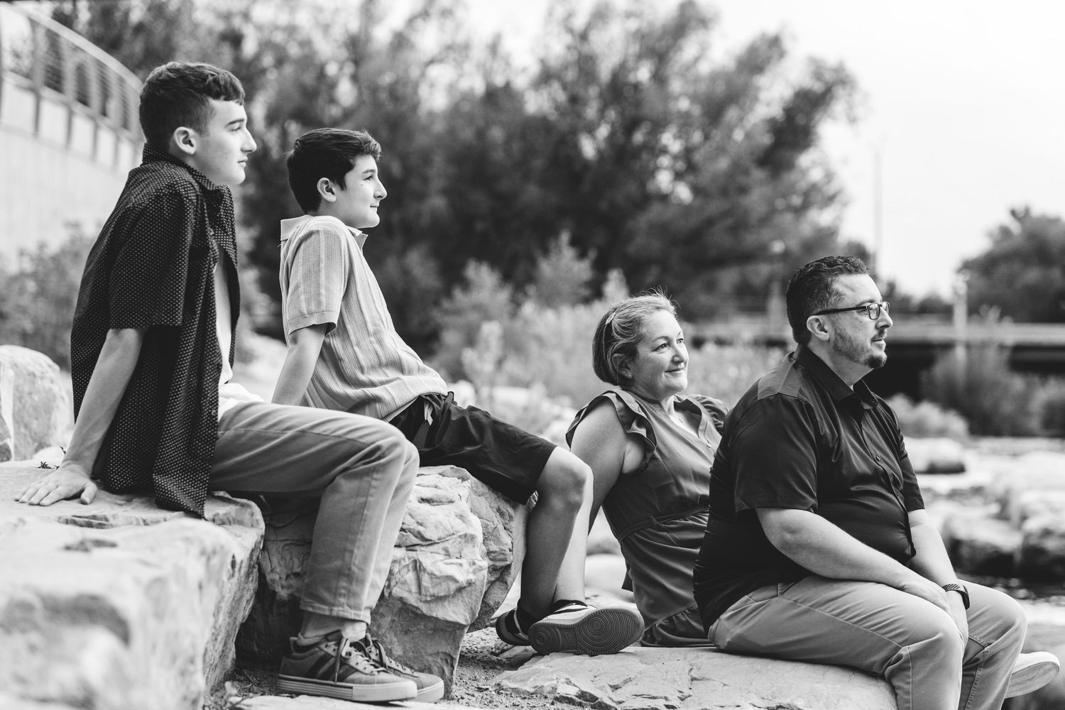 Serious black and white portrait of family of two teen boys and parents sitting on rocks overlooking Cache la Poudre River.