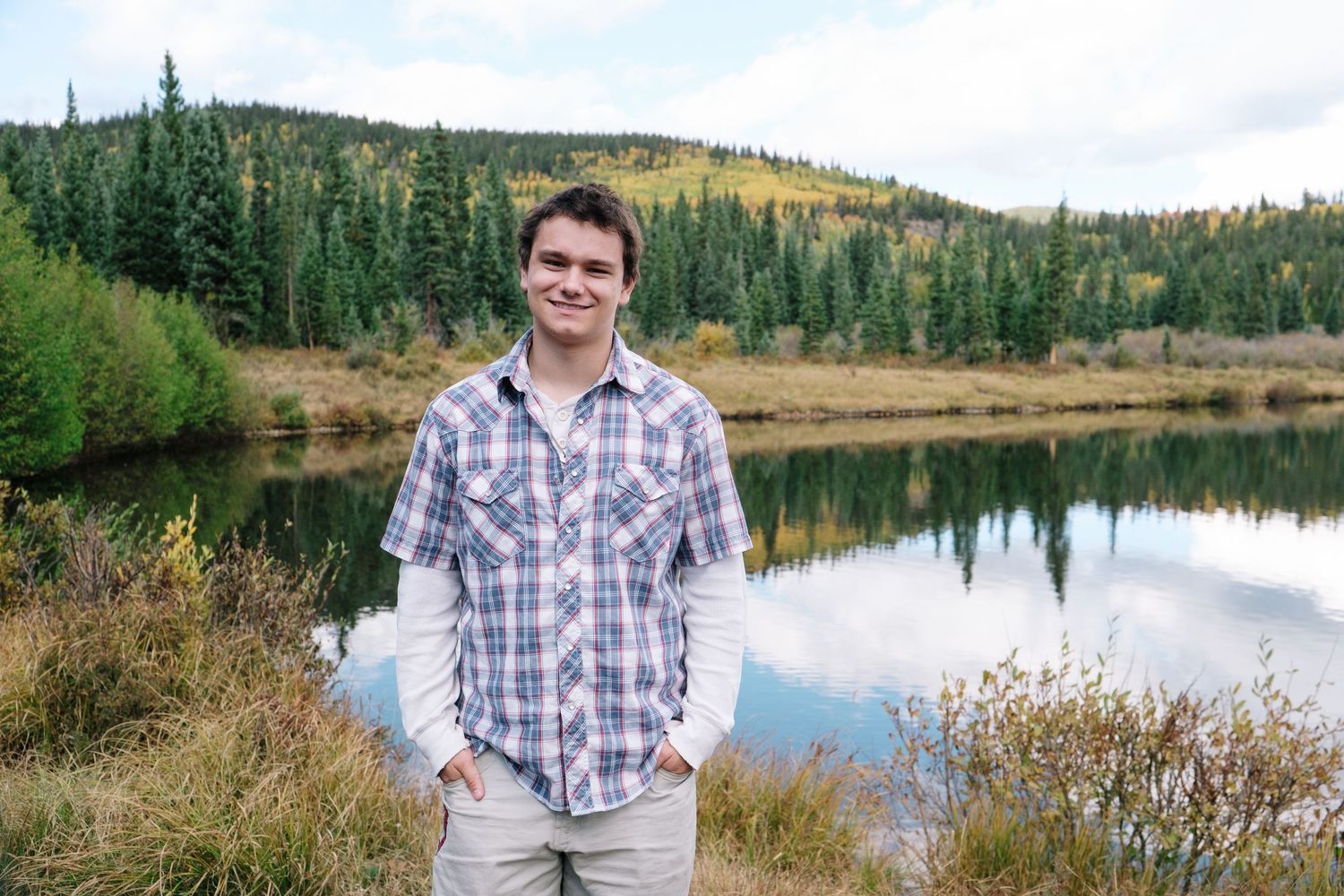Teen boy smiling, lake in background reflecting evergreen trees and blue skies, fall foliage in the fair background.
