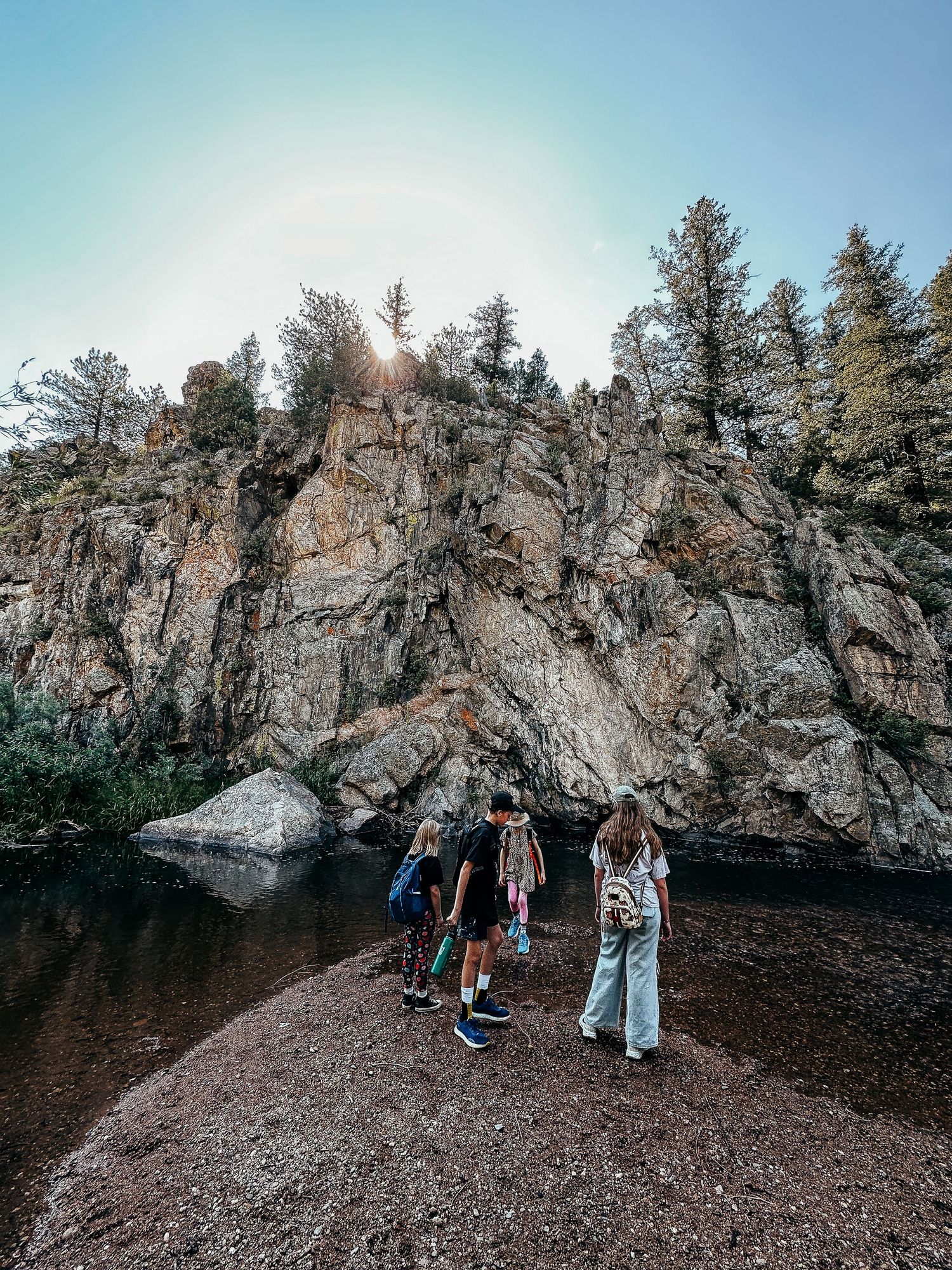 Wide angle shot of children standing on pebbly riverbank with large rock formation on other side of river.
