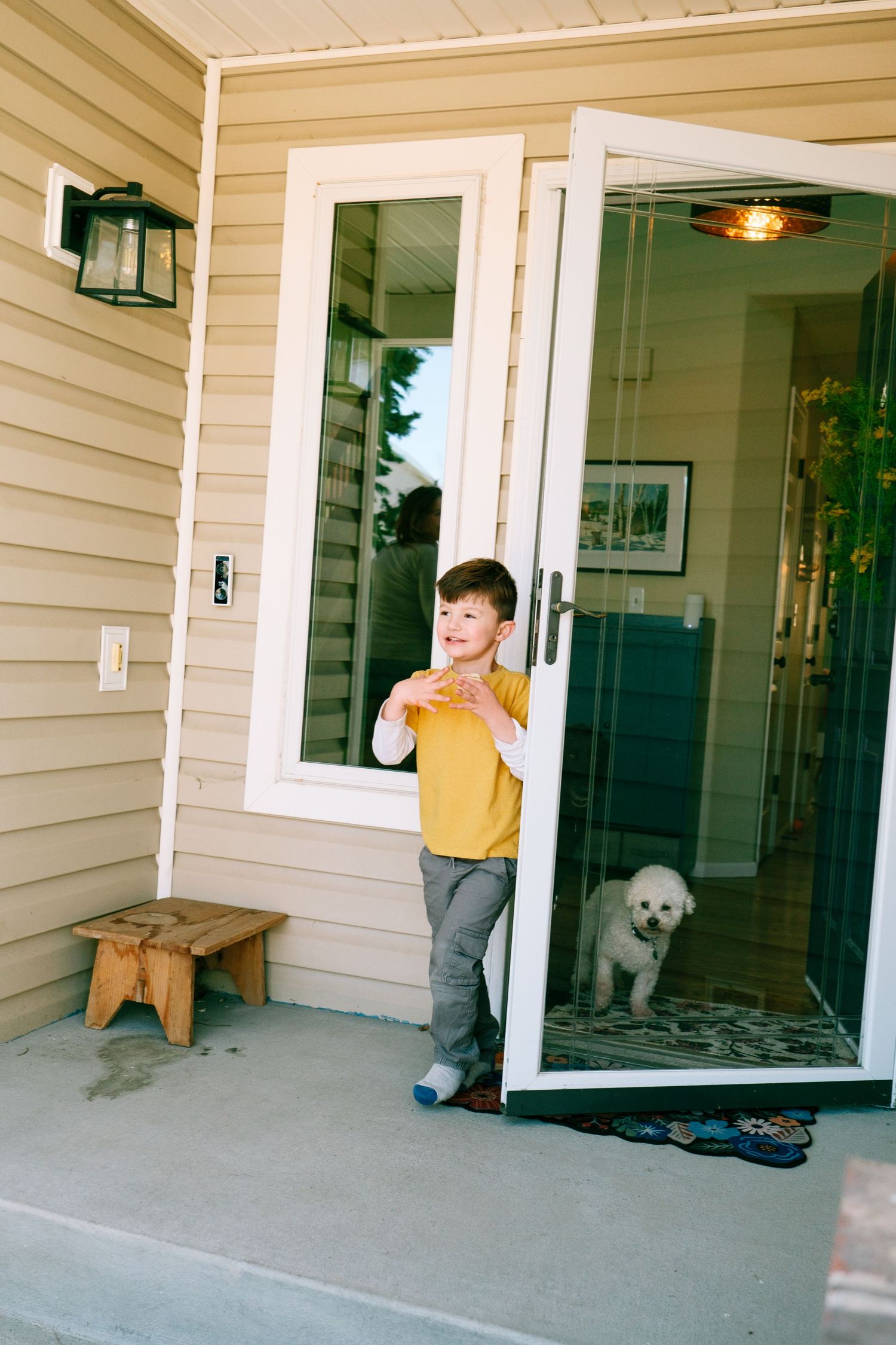 Young boy walking out of front door, small white dog behind the glass front door.
