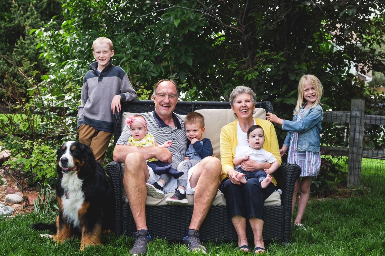 Grandparents sitting on outdoor couch with three grandsons, two granddaughters and Bernese Mountain dog, all smiling at the camera.