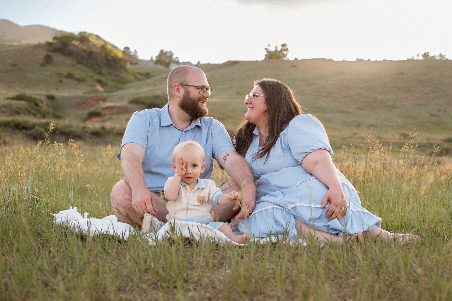 Mother, father, and son sitting in green grass around sunset at South Valley Park in Littleton, Colorado.  Toddler rubbing his eye, tiredly.
