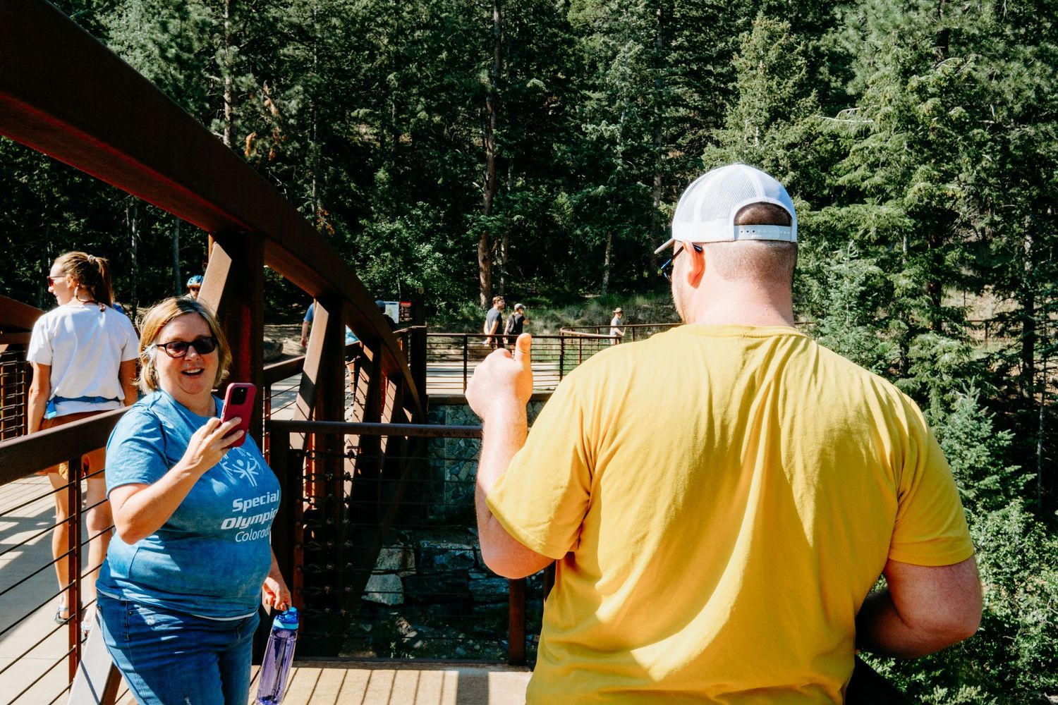 Mother taking picture of adult son standing on bridge, son giving a  thumbs up to her cell phone camera on group hike.
