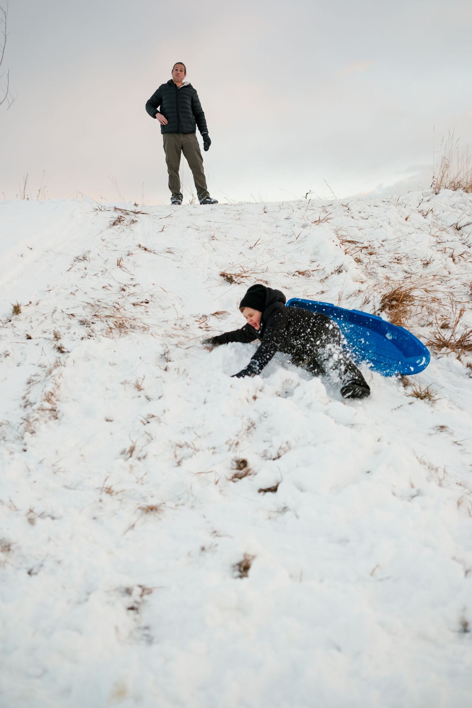 Family sledding photo session, father standing on hill, child crashing in snow on blue sled.
