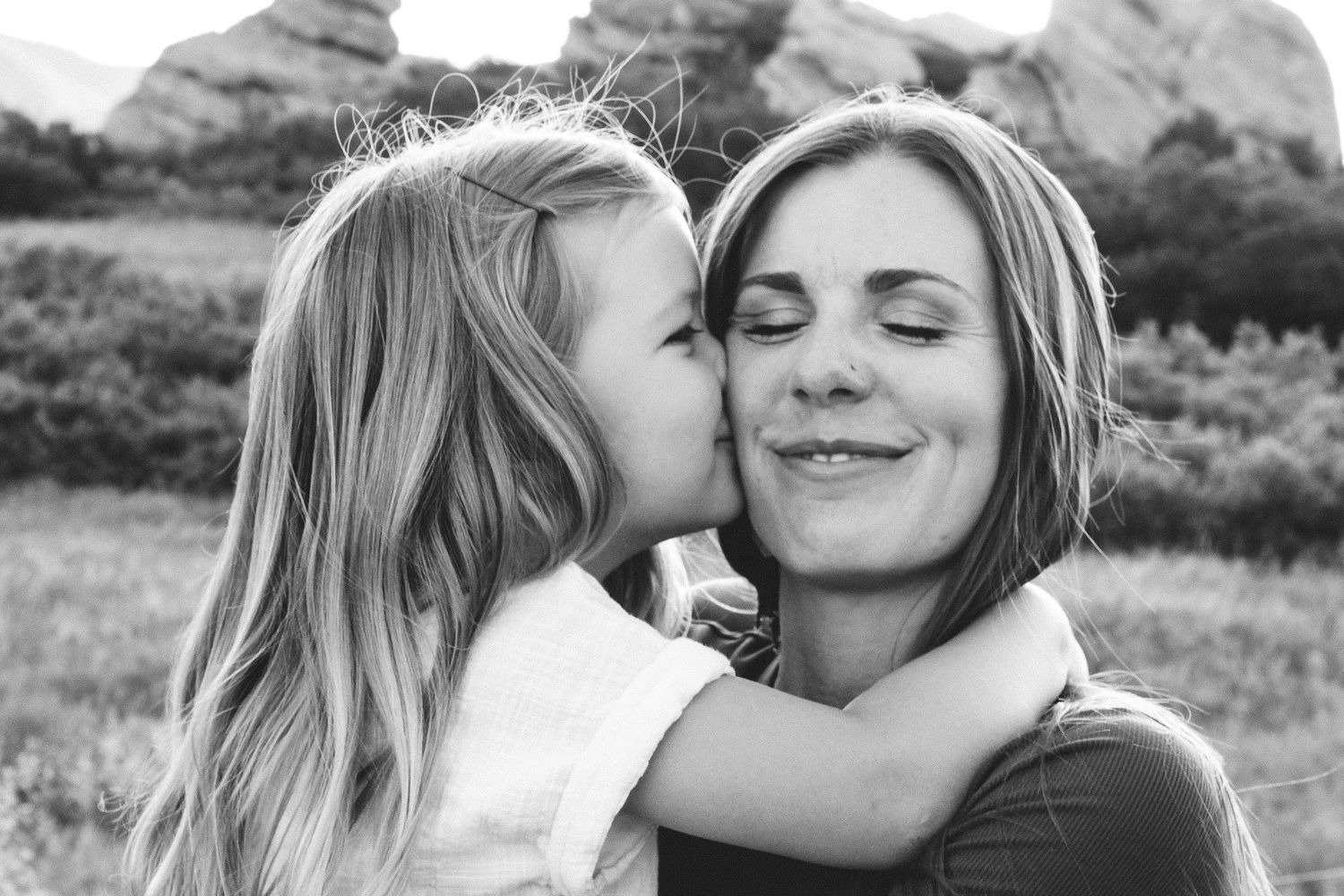 Black and white photo of young girl kissing mother on the cheek, mother smiling with eyes closed.