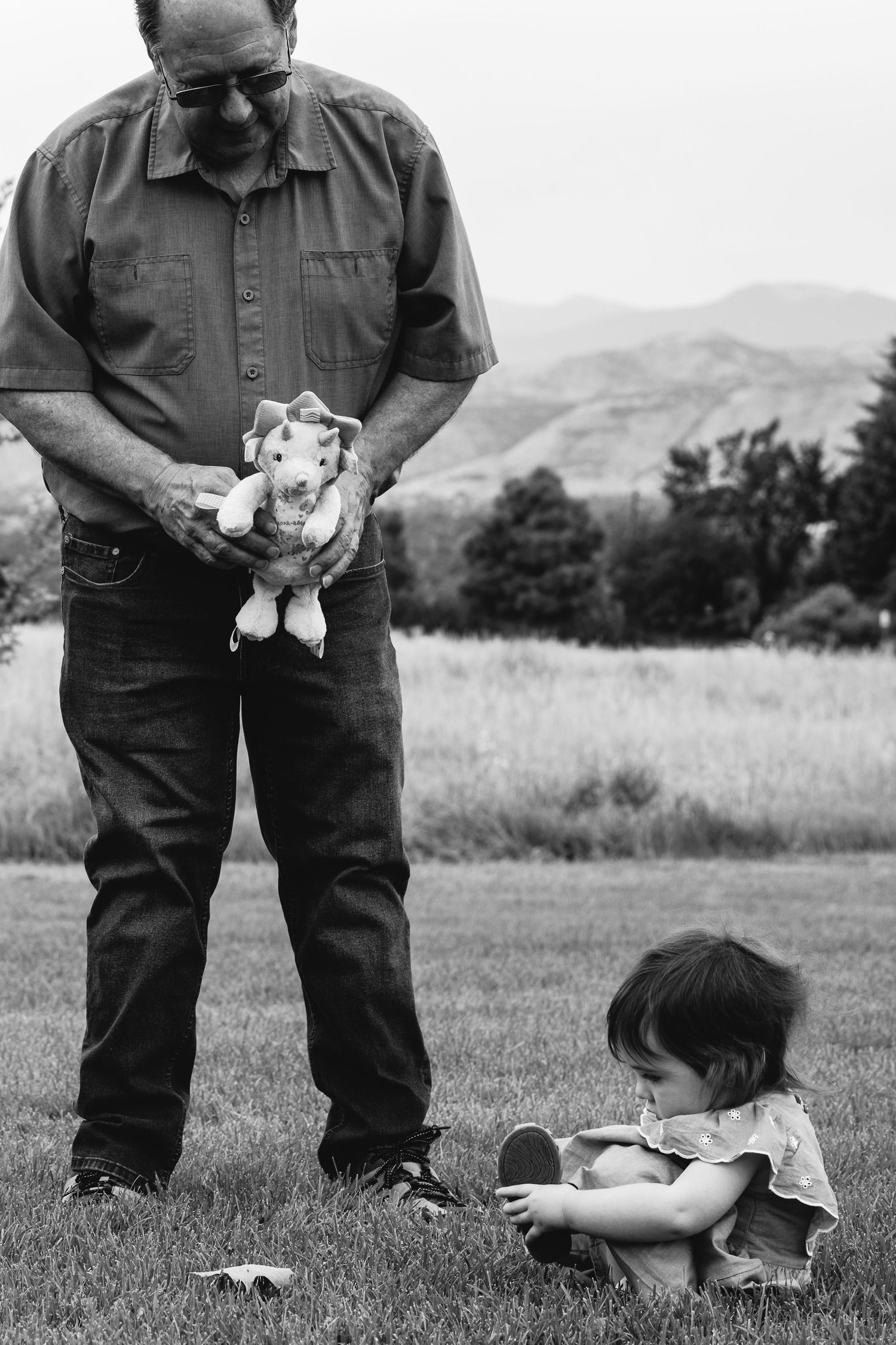 Black and white candid of sweet grandpa holding stuffed animal out for toddler girl who is sitting on grass.
