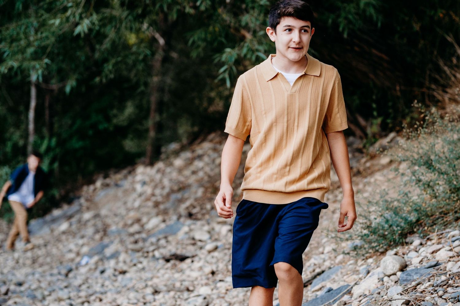 Teen boy walking on rocky bank of Cache la Poudre, brother following behind him.