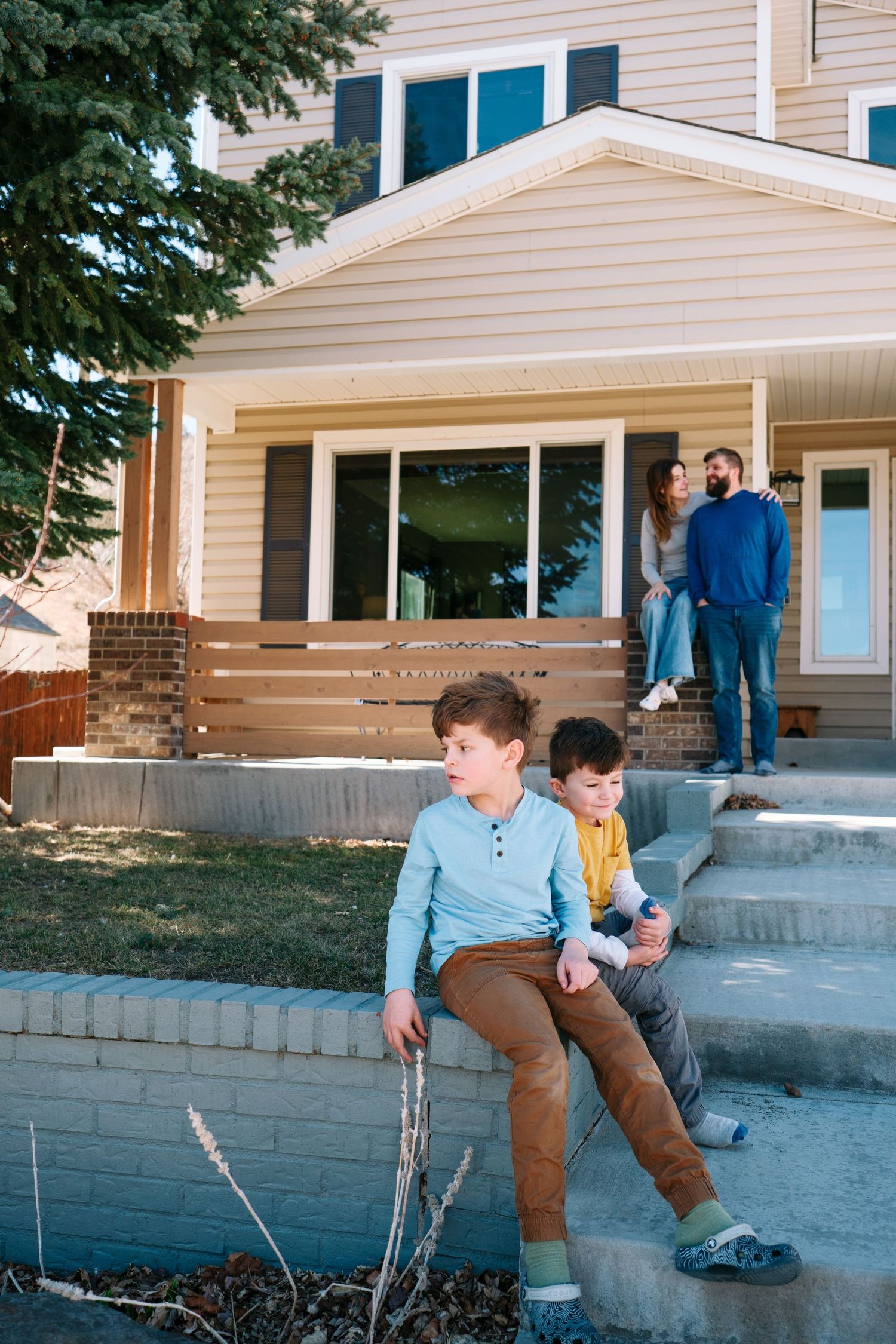 Two brothers sitting on retaining wall in front yard, mother and father sitting on the porch in the background.