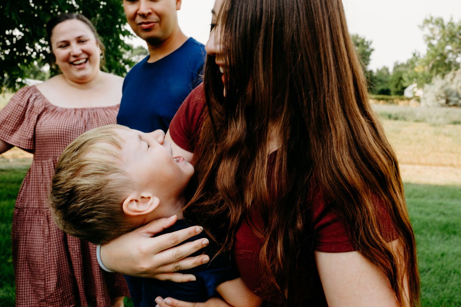 Big sister playfully holding younger brother, brother laughing, parents blurred in background.