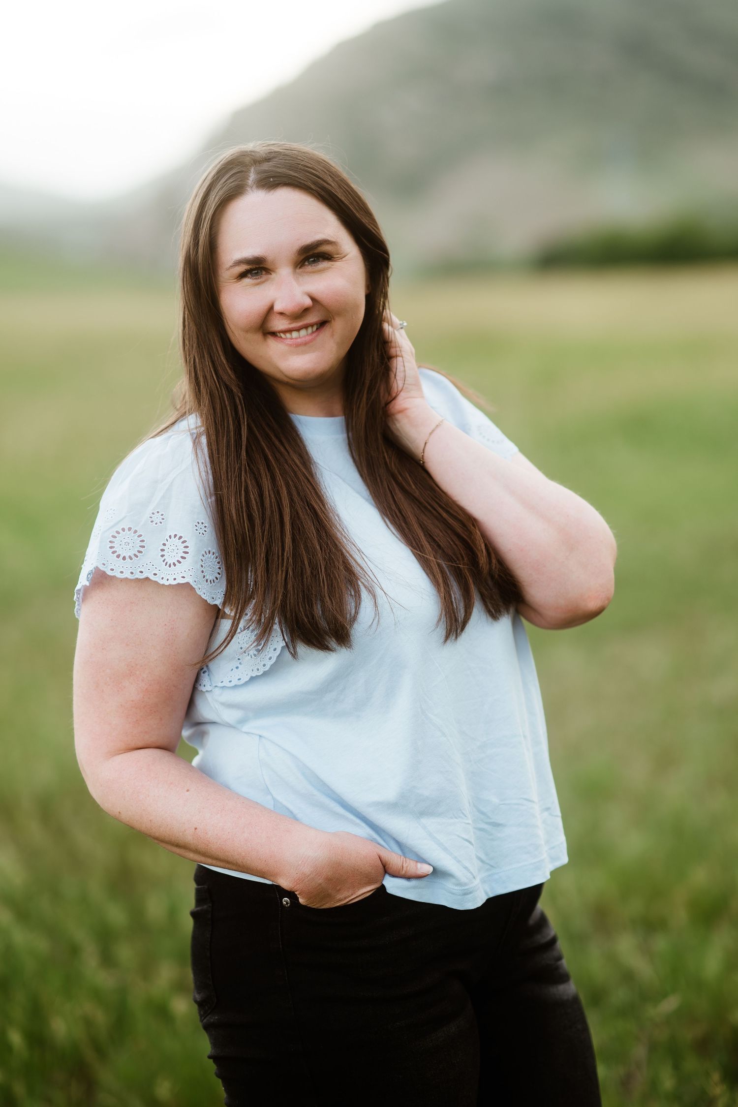 Headshot of Maggie Theis, owner of Maggie Theis Photography.  Woman standing in green meadow, foothills in background, wearing blue shirt.
