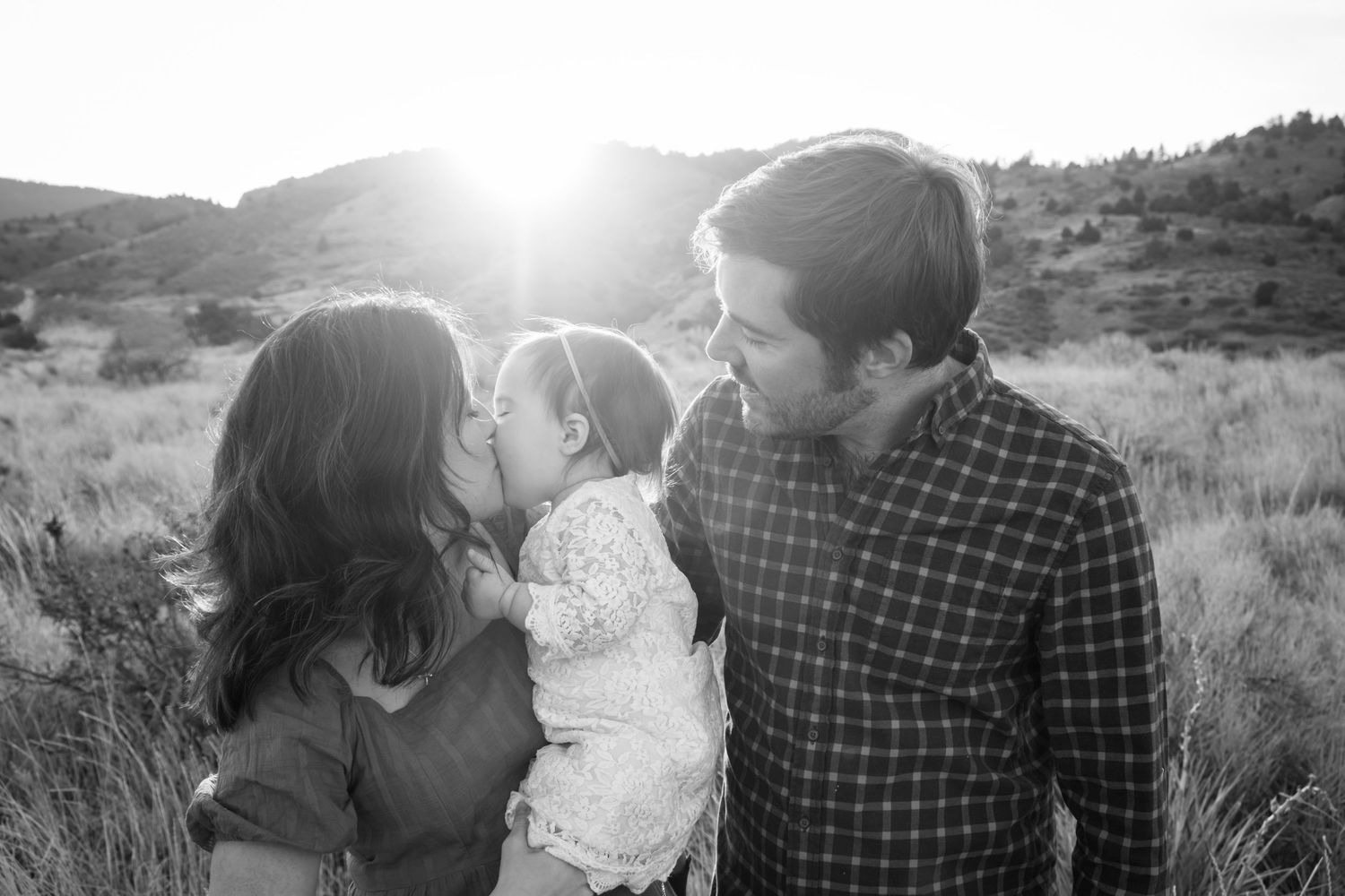 Mother, father and toddler daughter snuggling close together at Mount Falcon East in Morrison, Colorado, sun bursting over foothills.