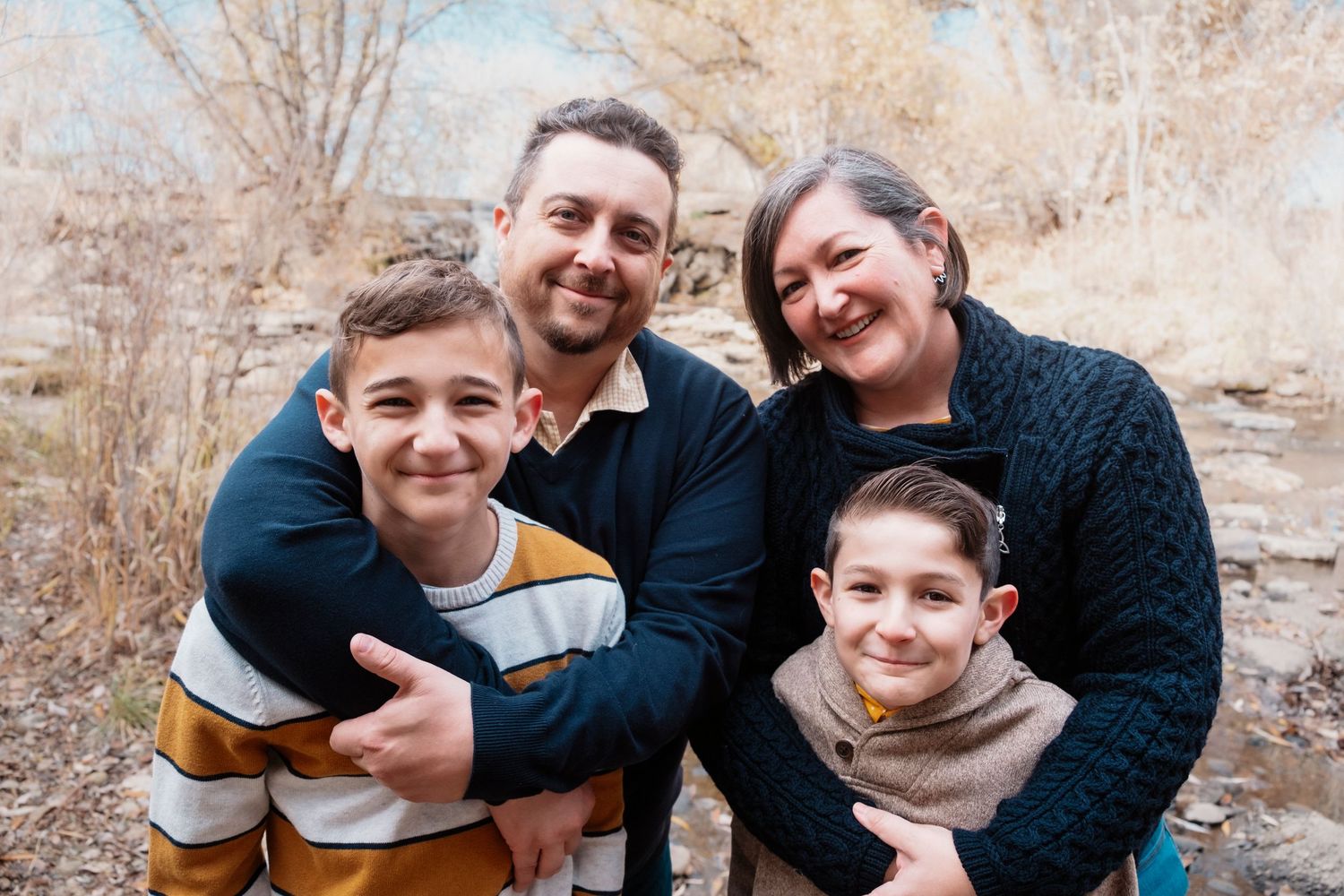Father and mother standing, two sons standing in front of them, all smiling and snuggling together, fall foliage in background.