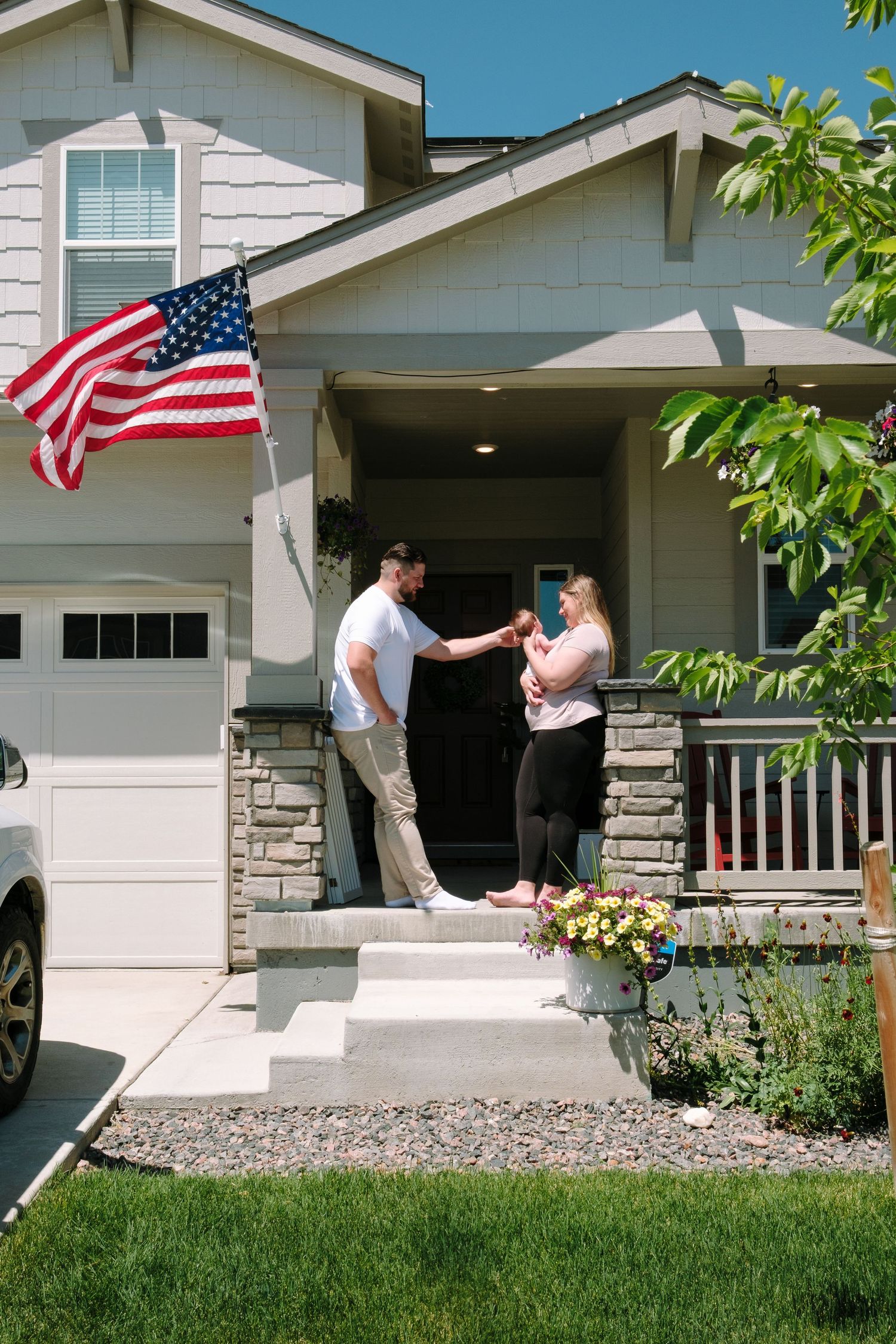 Mother and father standing on porch, each leaning against posts, American flag flapping in wind.