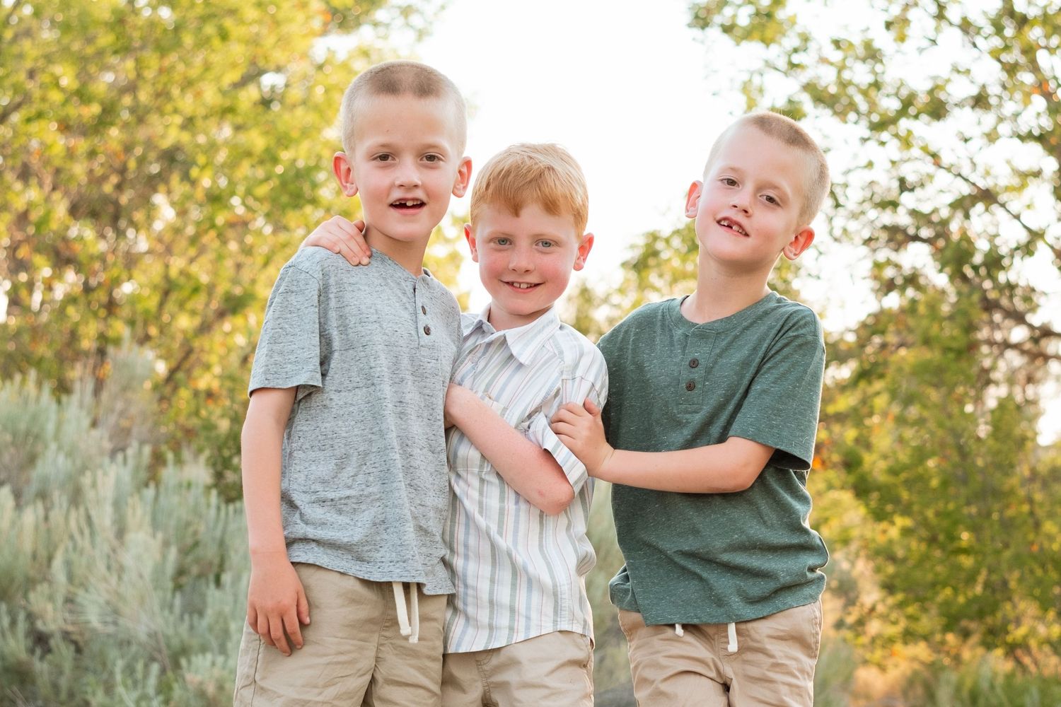 Three boy cousins standing closely together at a park, all smiling.