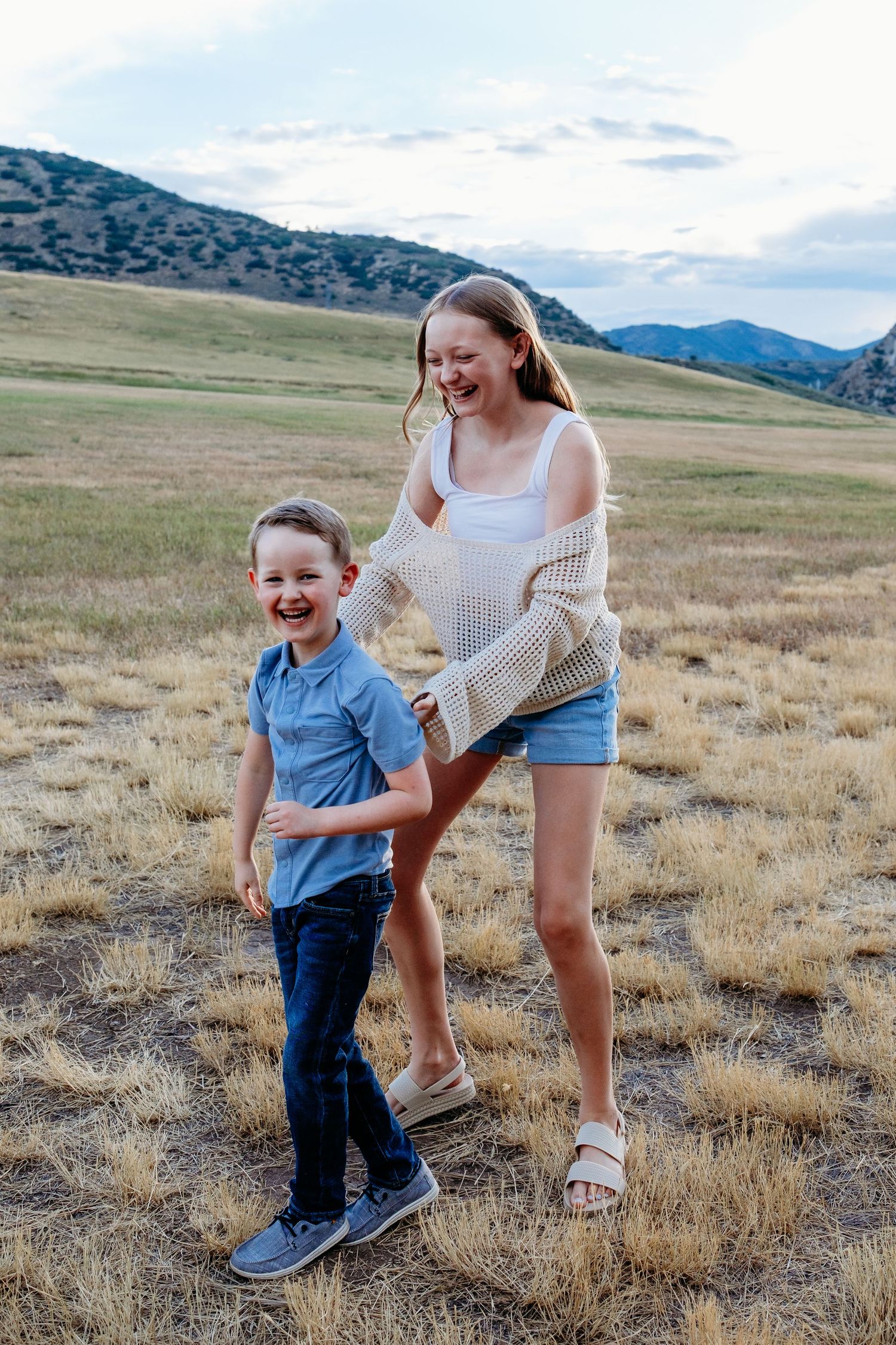 Brother and sister laughing playfully in field at Hildebrand park with blue skies and foothills in background.