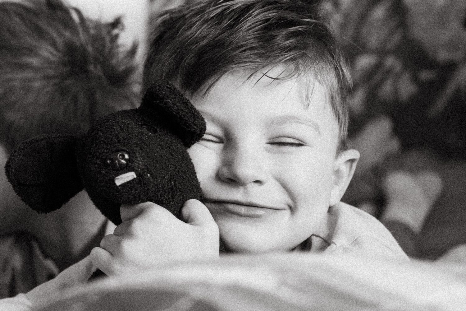 Black and white portrait of young boy smiling with eyes closed, hugging a black stuffed dog during documentary family photo session.