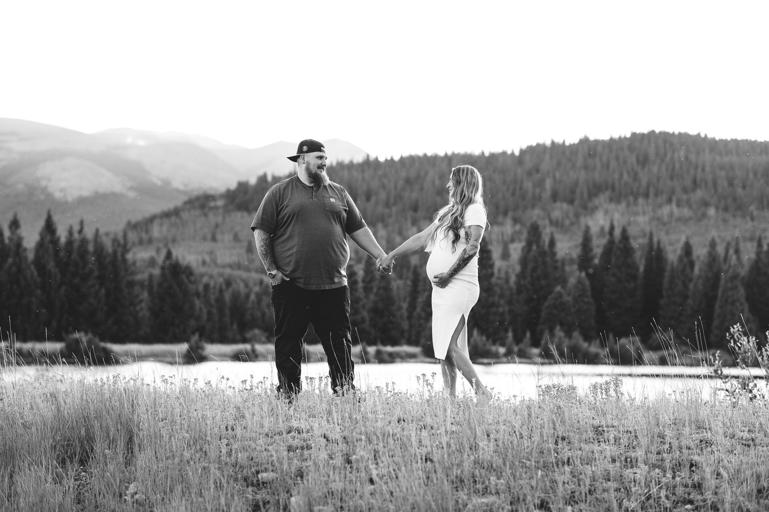 Husband and expecting wife holding hands, looking at one another, lake, trees and mountains in background.