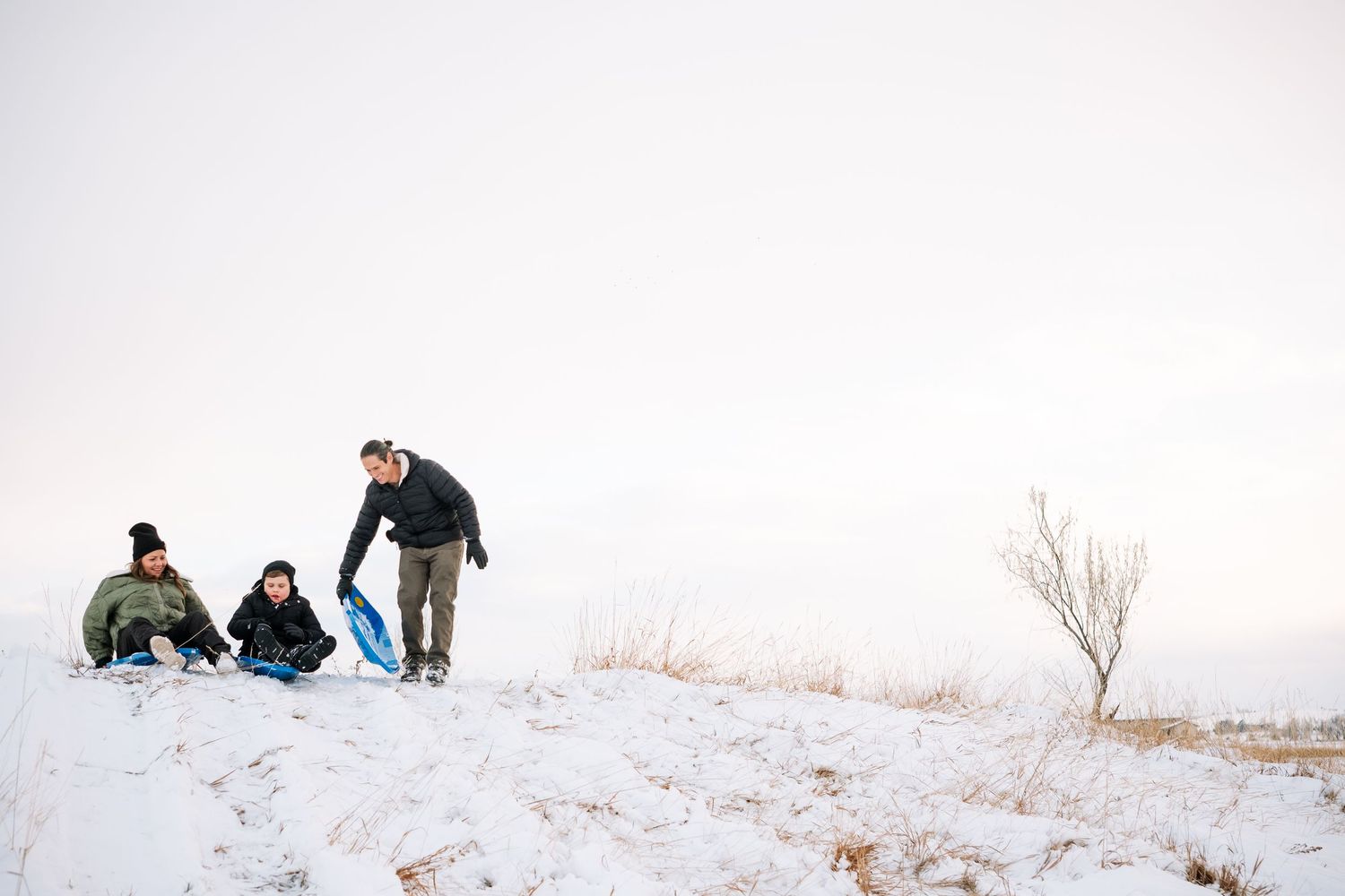 Mother, son and father preparing to sled down a snowy hill on blue sleds.