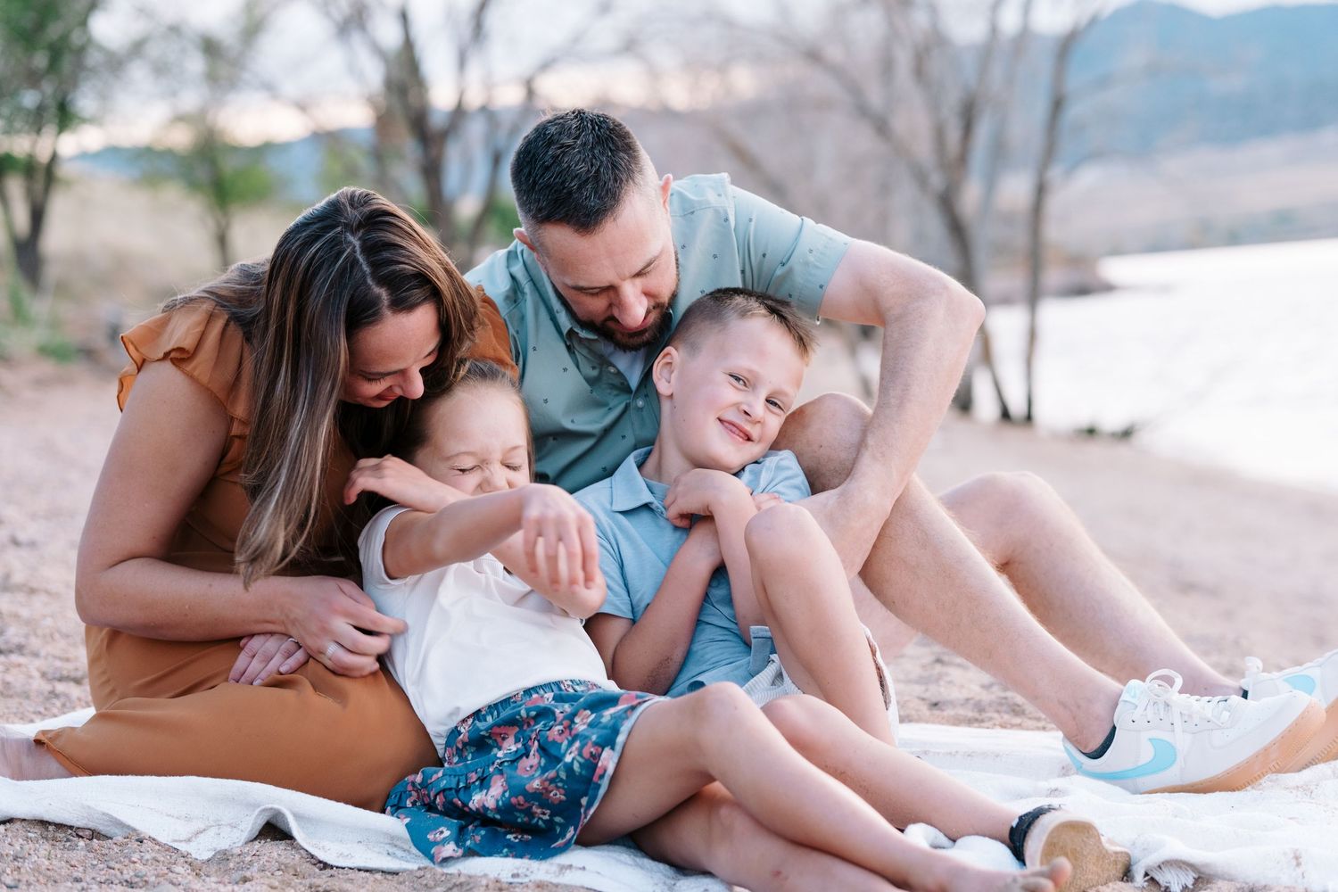Family of four sitting on white blanket, parents playfully tickling kids.