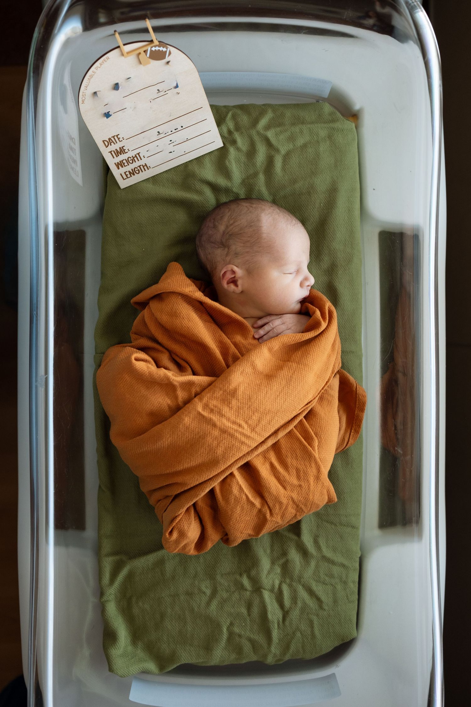 Overhead photo of baby boy swaddled in rusty orange muslim swaddle, sleeping peacefully in bassinet at hospital