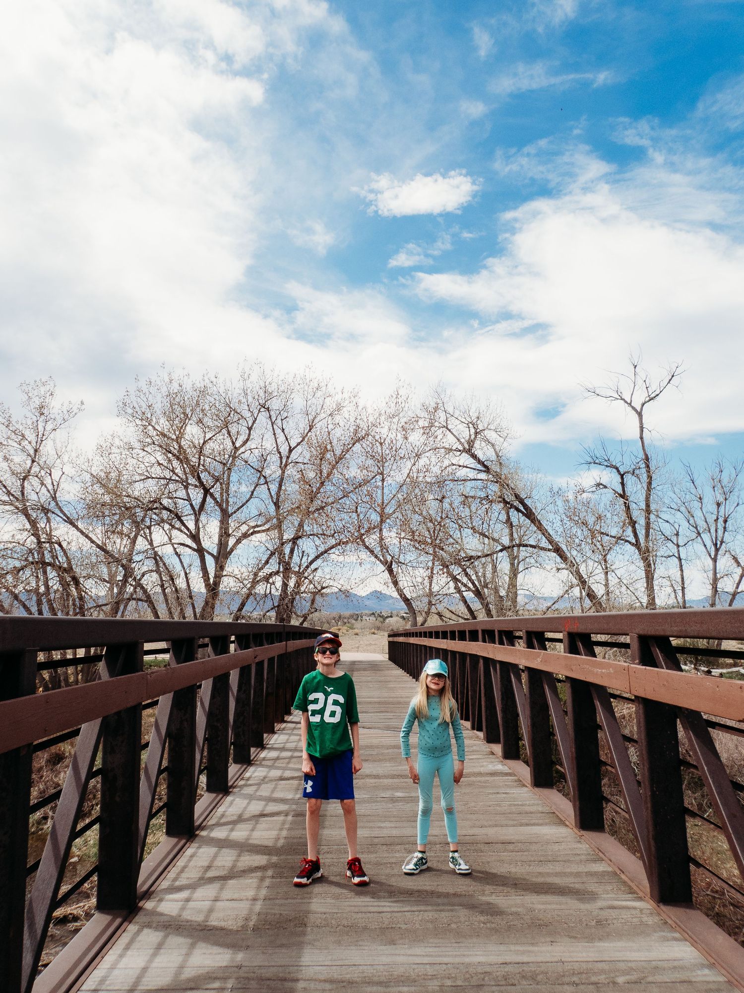 Brother and sister standing on bridge, trees, clouds and blue skhy in background.
