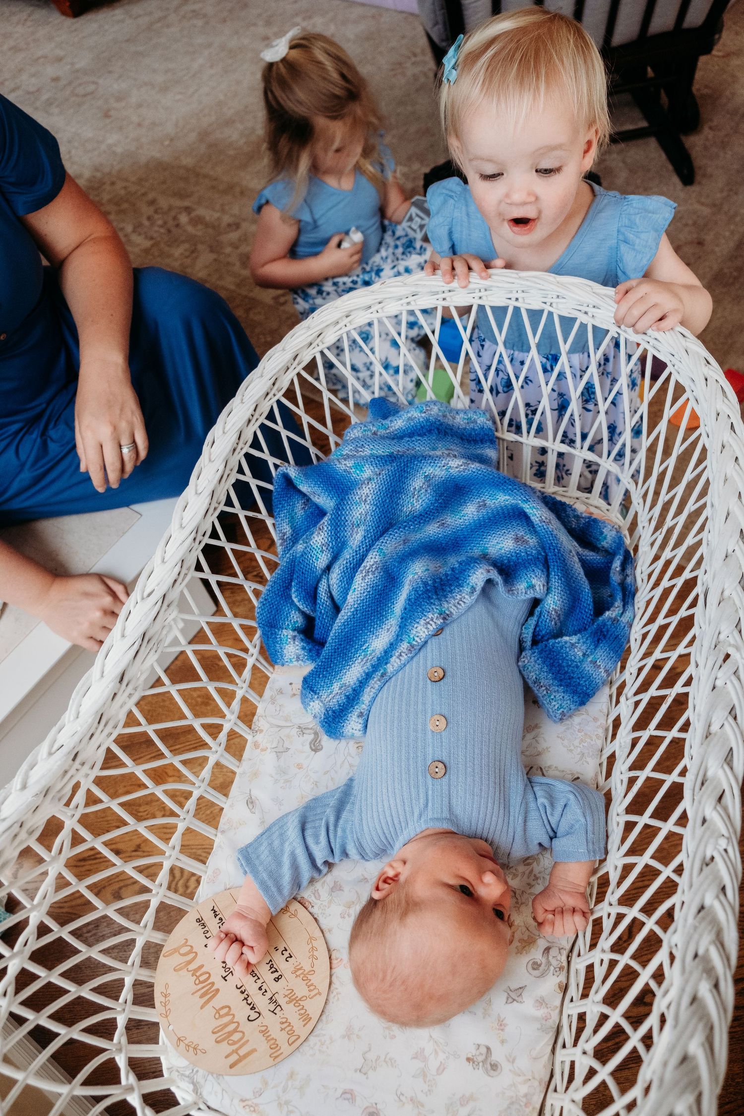 Baby brother resting in bassinet as toddler sister looks toward him with face of wonder.