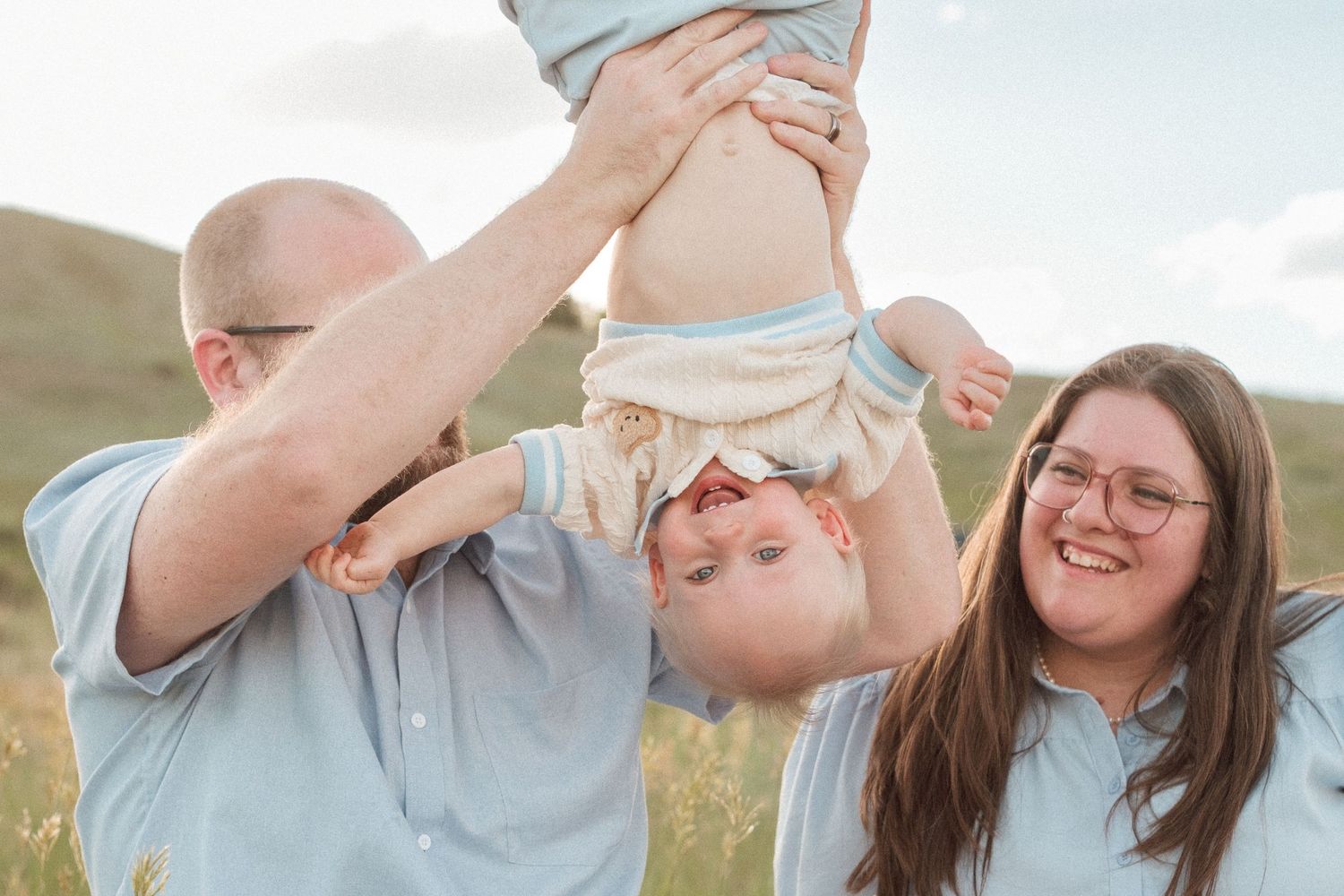 Mother and father sitting in green grasses, father holding one year old son upside down, son laughing.