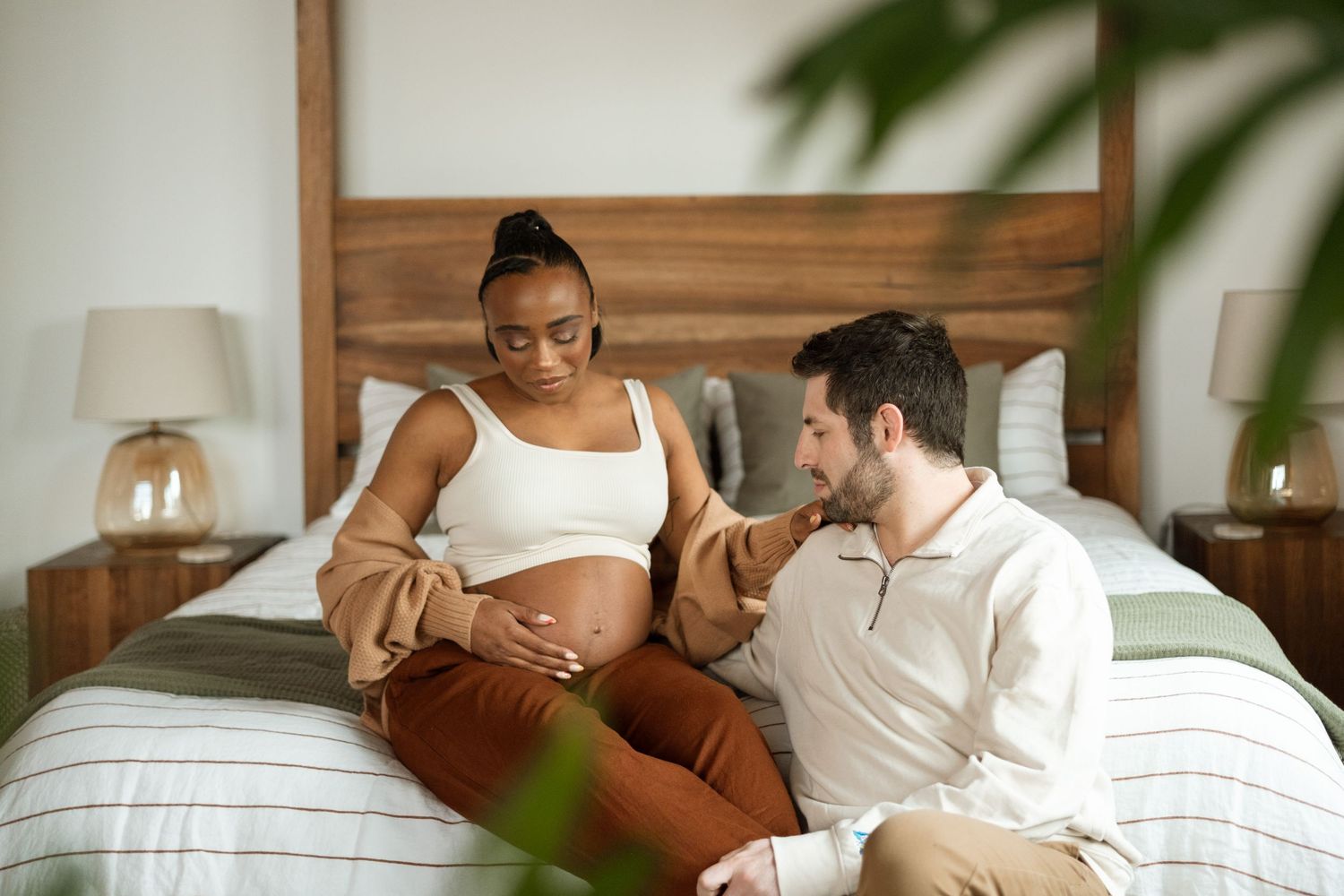 Pregnant couple sitting on bed framed by monstera leaves.