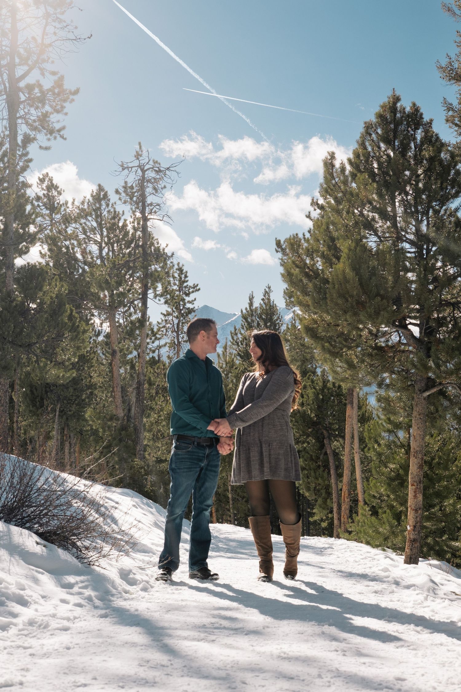 Engaged couple holding hands and looking at one another on snowy trail at Sapphire Point Overlook, surrounded by tall trees.