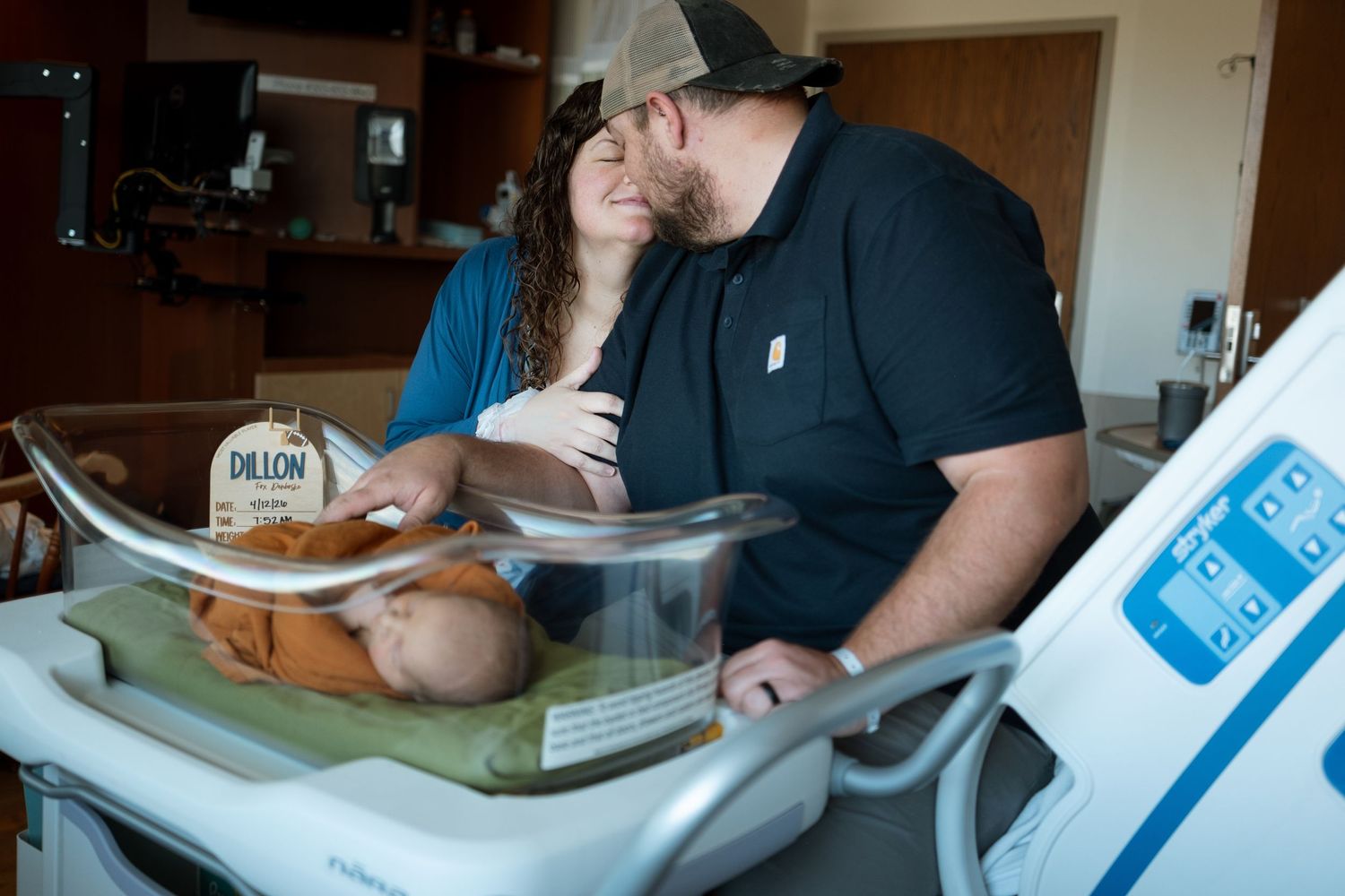 New mother and father snuggling closely on hospital bed, newborn baby in bassinet in hospital room.