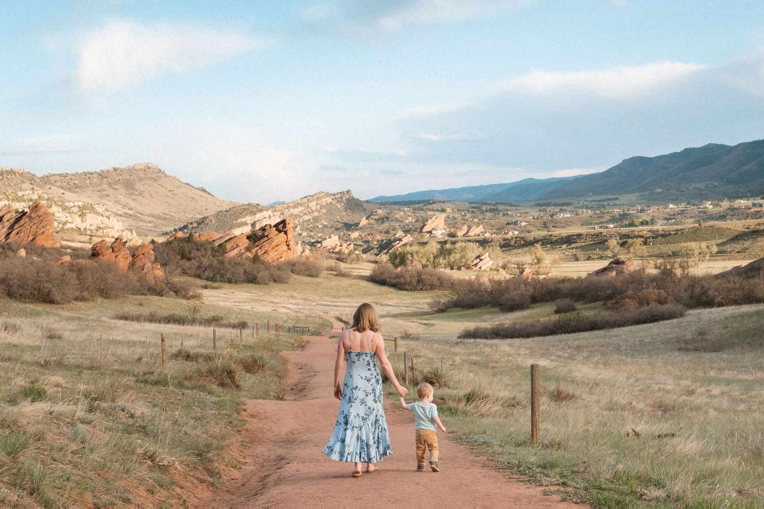 Landscape portrait of mother and toddler walking away, toward red rocks of South Valley Park in Littleton, Colorado.