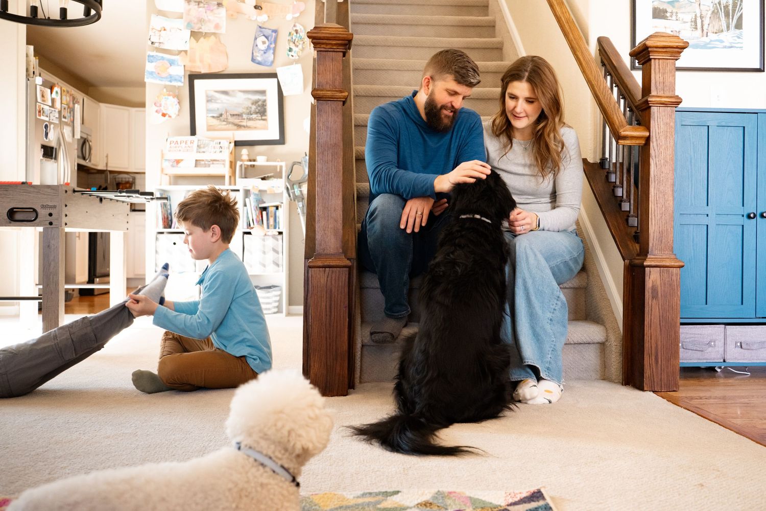 In home, documentary photo of parents sitting on stairs petting black dog, child aged son playing with toddler aged brother on the floor.