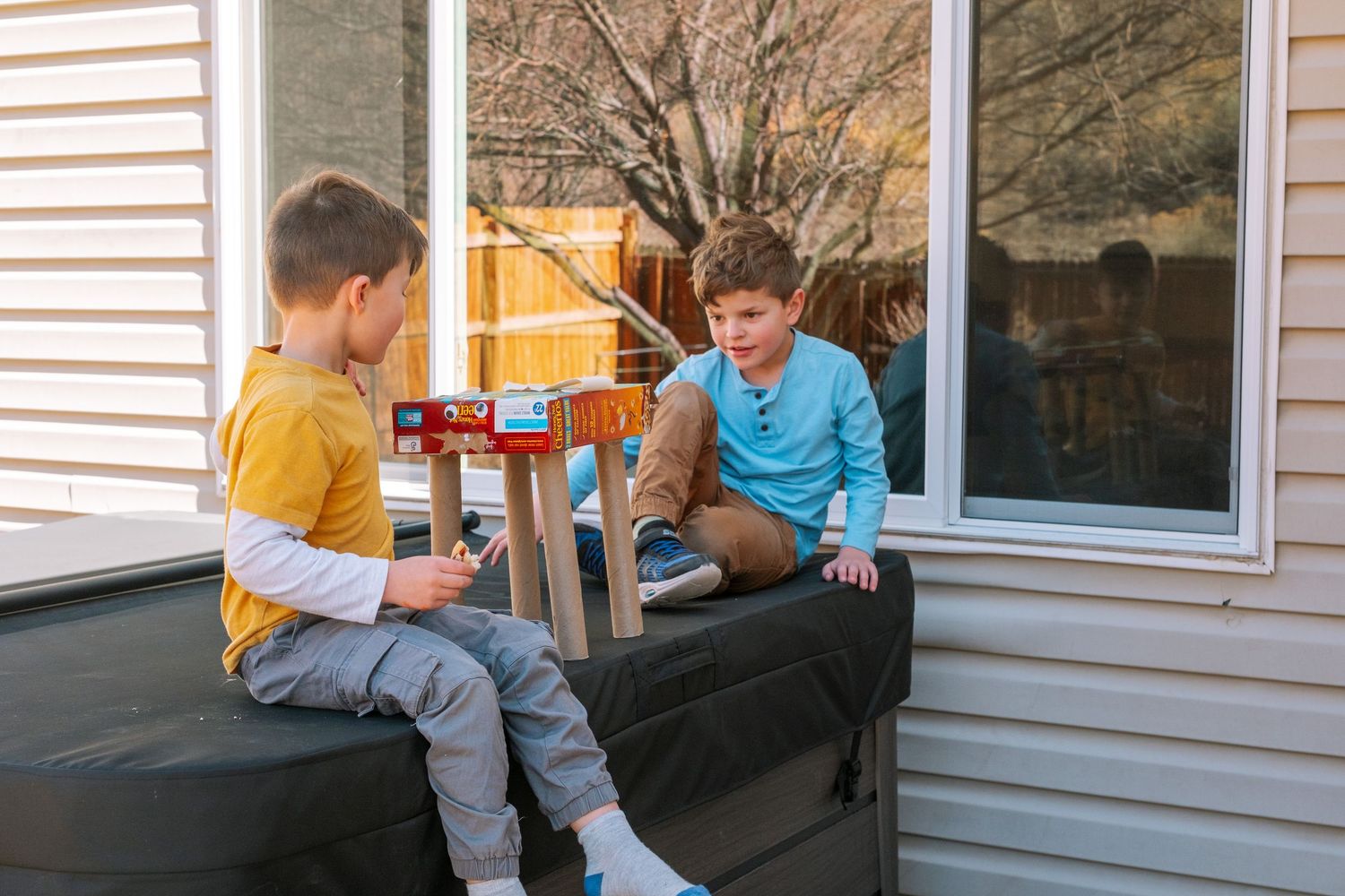 Brothers sitting on top of closed hot tub with cereal box robot creation.