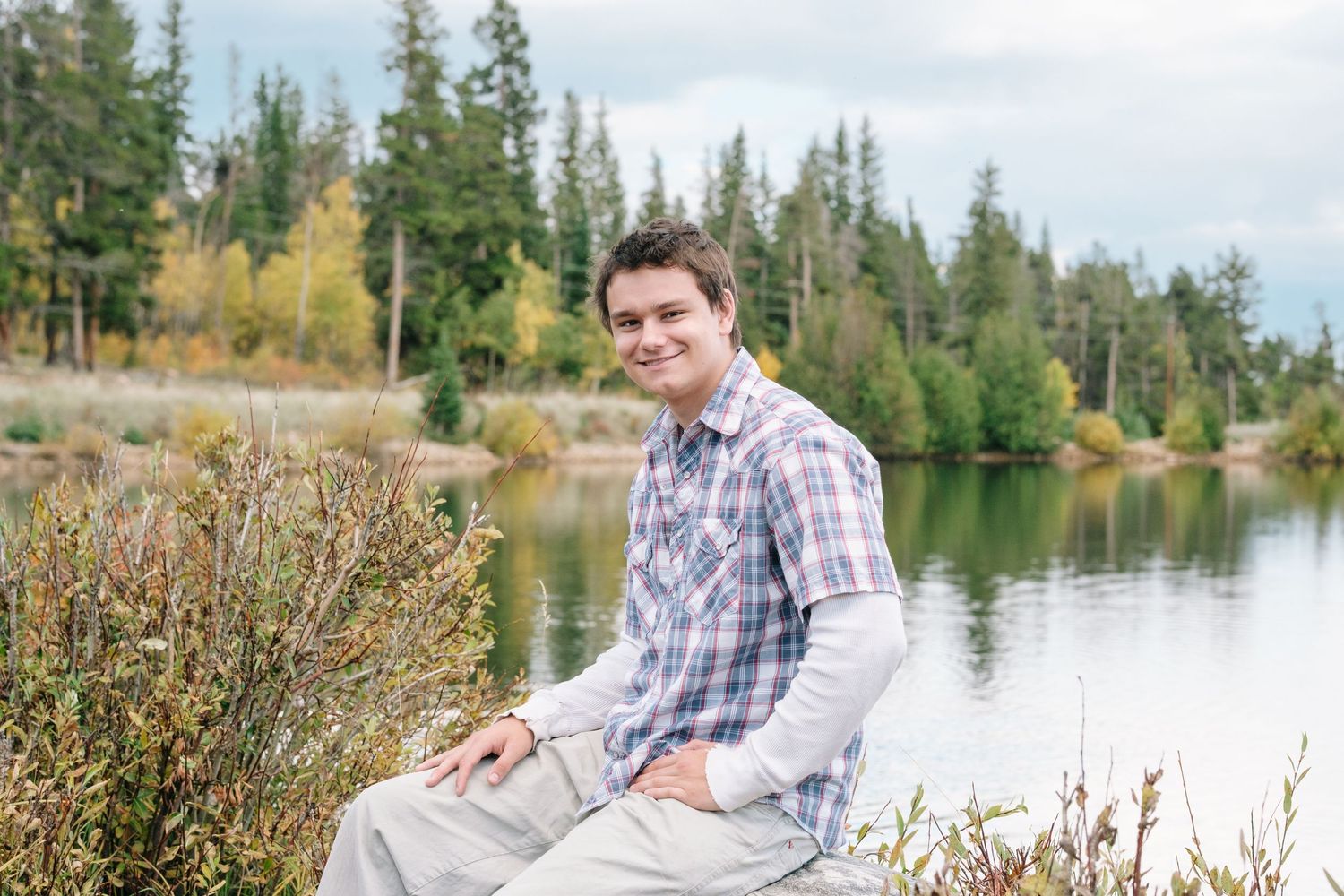 Teen boy sitting on boulder, lake and trees in the background.