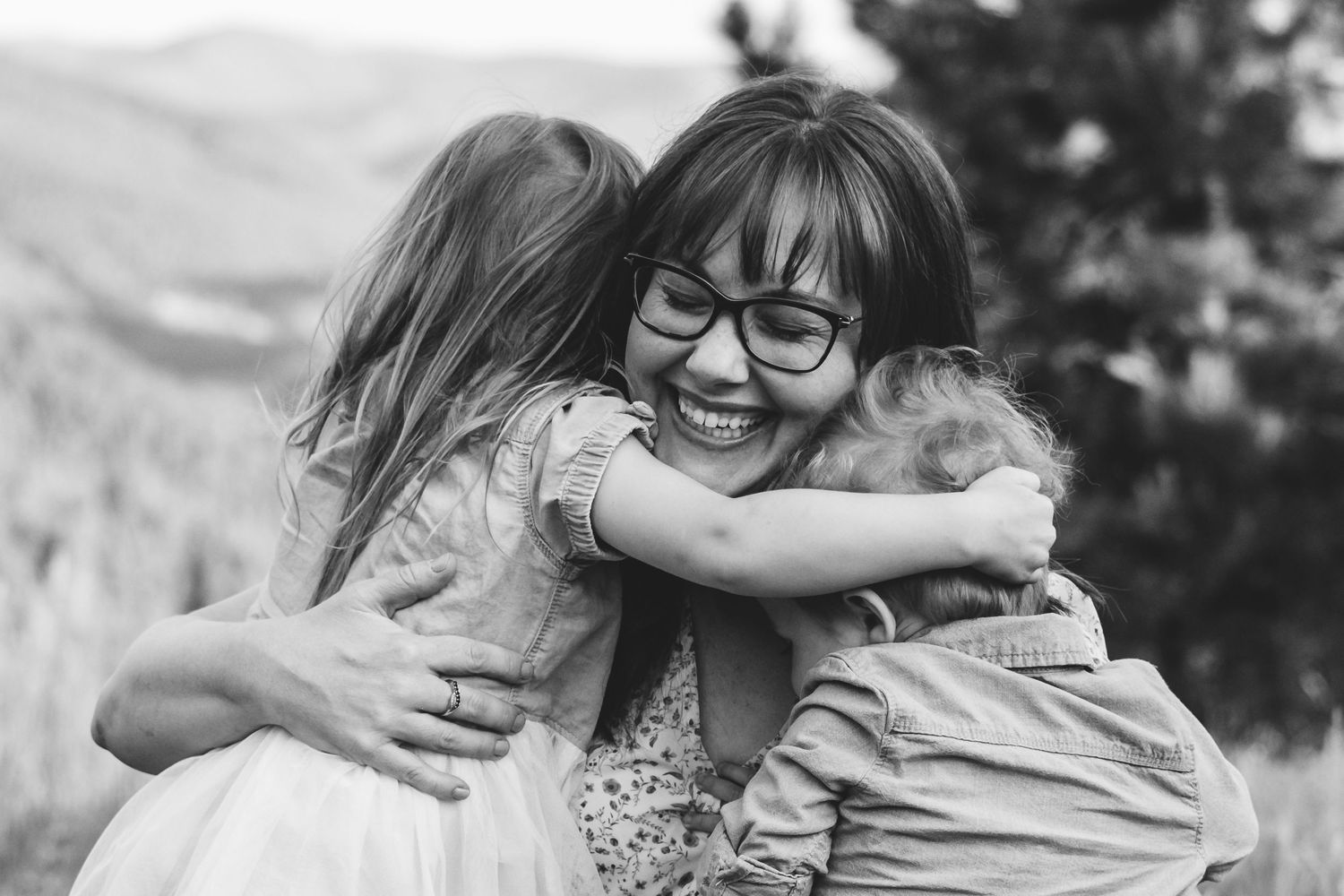 Black and white of mother as son and daughter wrap their arms around her, mother's eyes are closed, smiling widely.