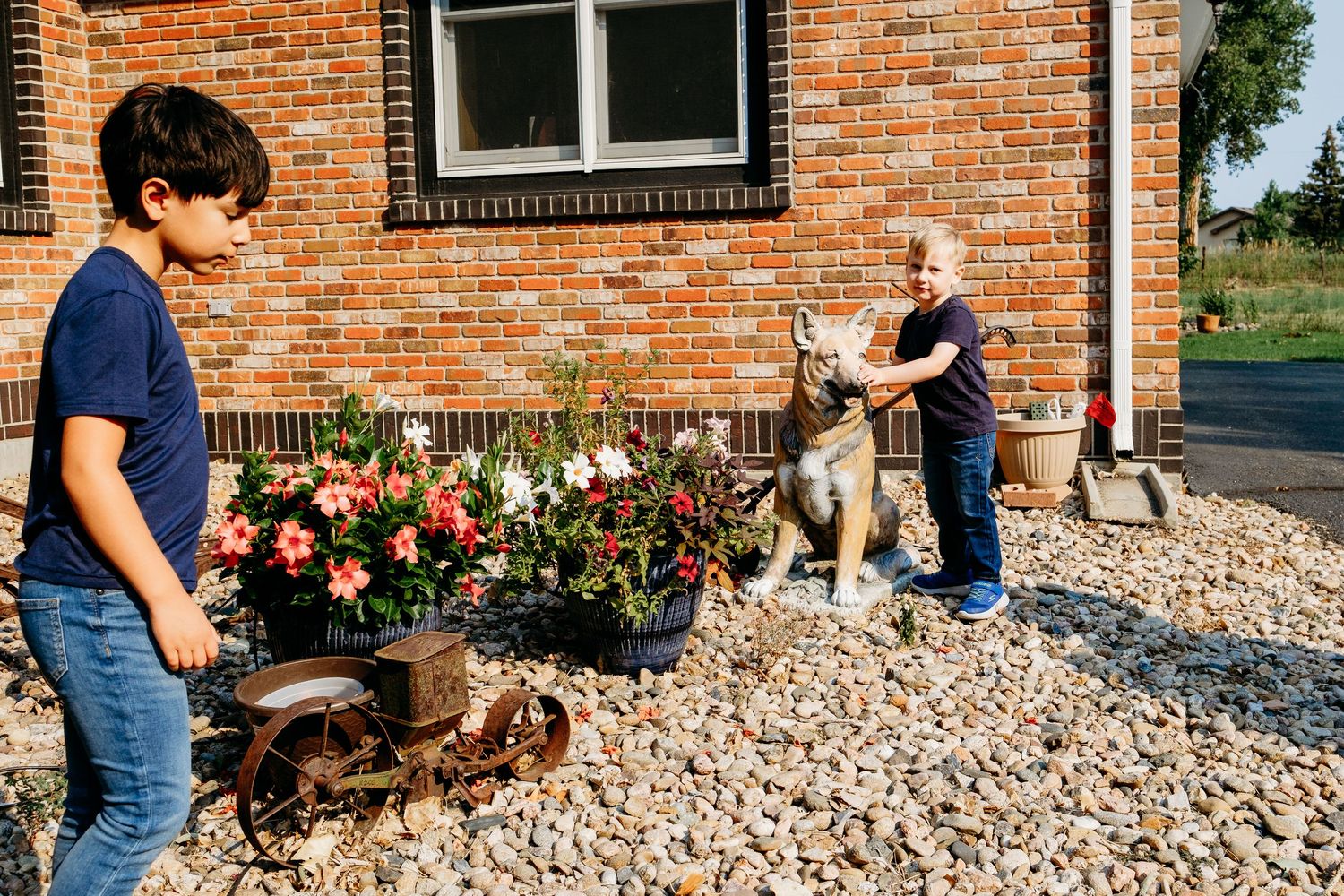 Child touching dog statue, brother standing nearby flowers.