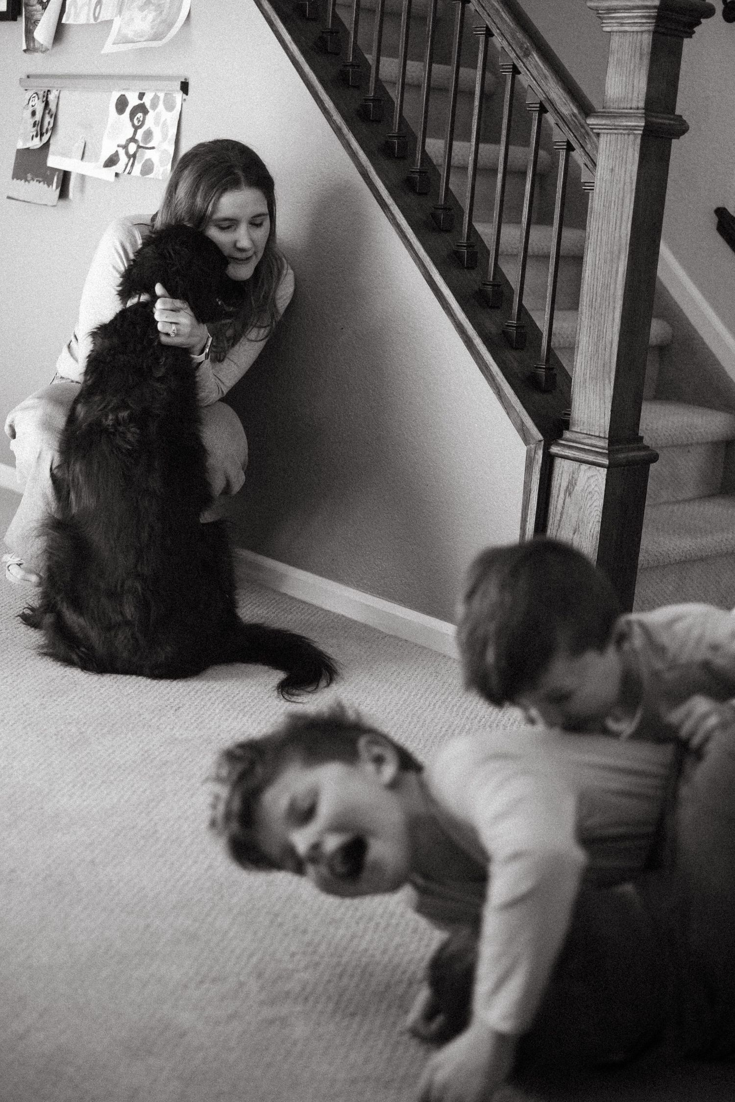 Black and white portrait of mother in the background snuggling large black dog, children in the foreground wrestling with dad, laughing.